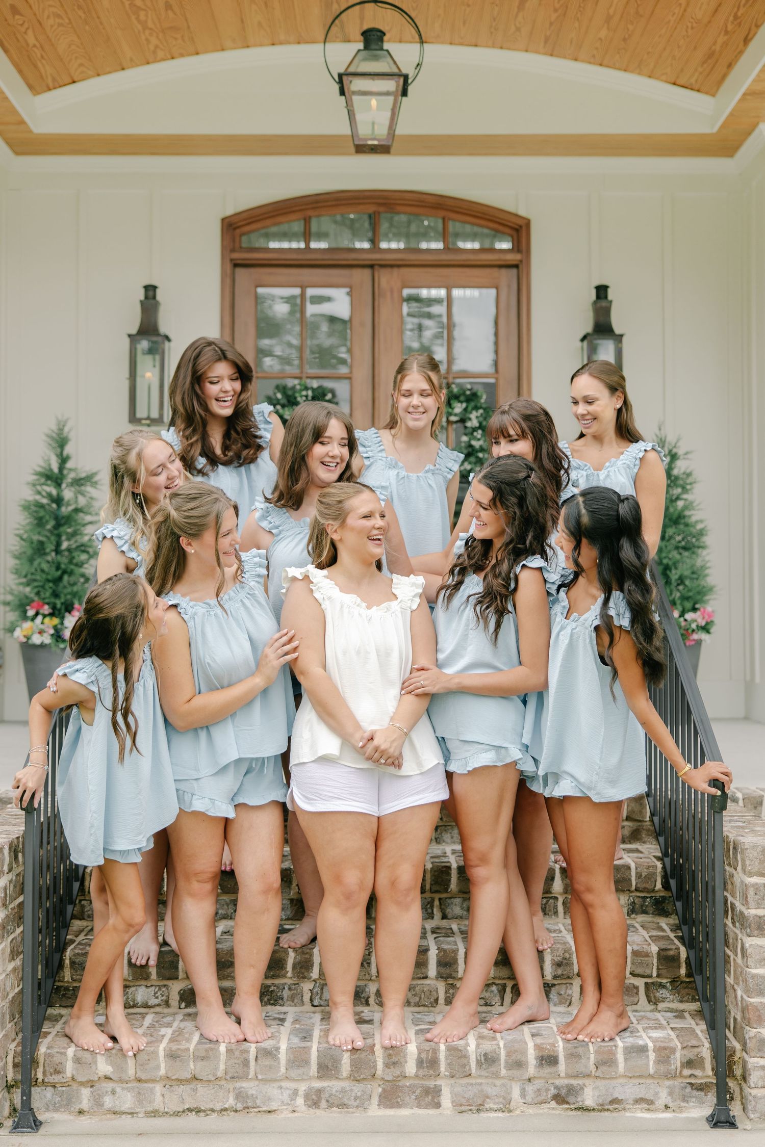 Bridesmaids in matching light blue rompers gather on brick steps laughing with bride in white before wedding.