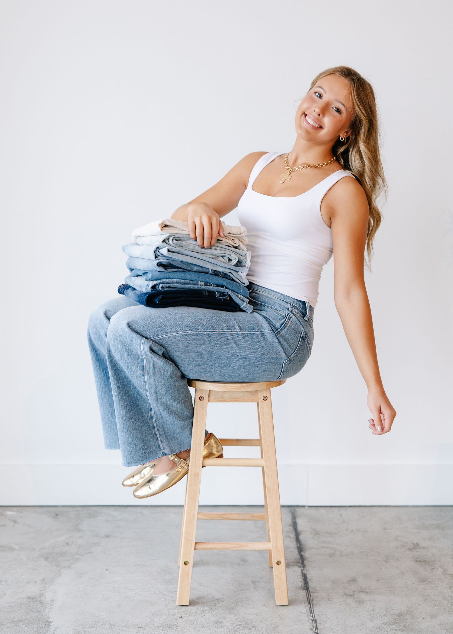 Woman sitting on a stool with a stack of folded denim jeans, wearing a white tank and wide-leg jeans.