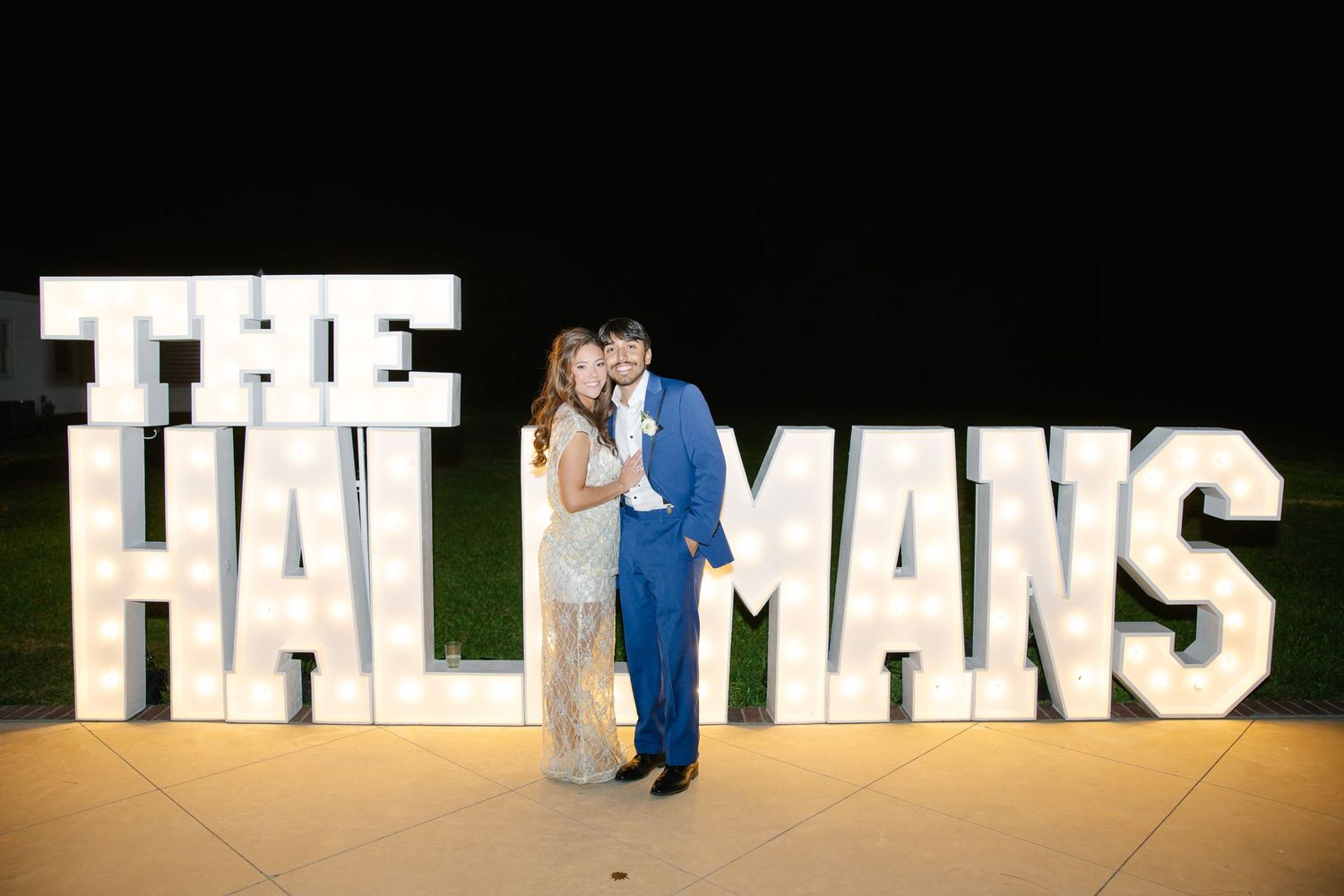 Couple kisses in front of illuminated marquee letters spelling THE HALLMANS at outdoor evening wedding reception.