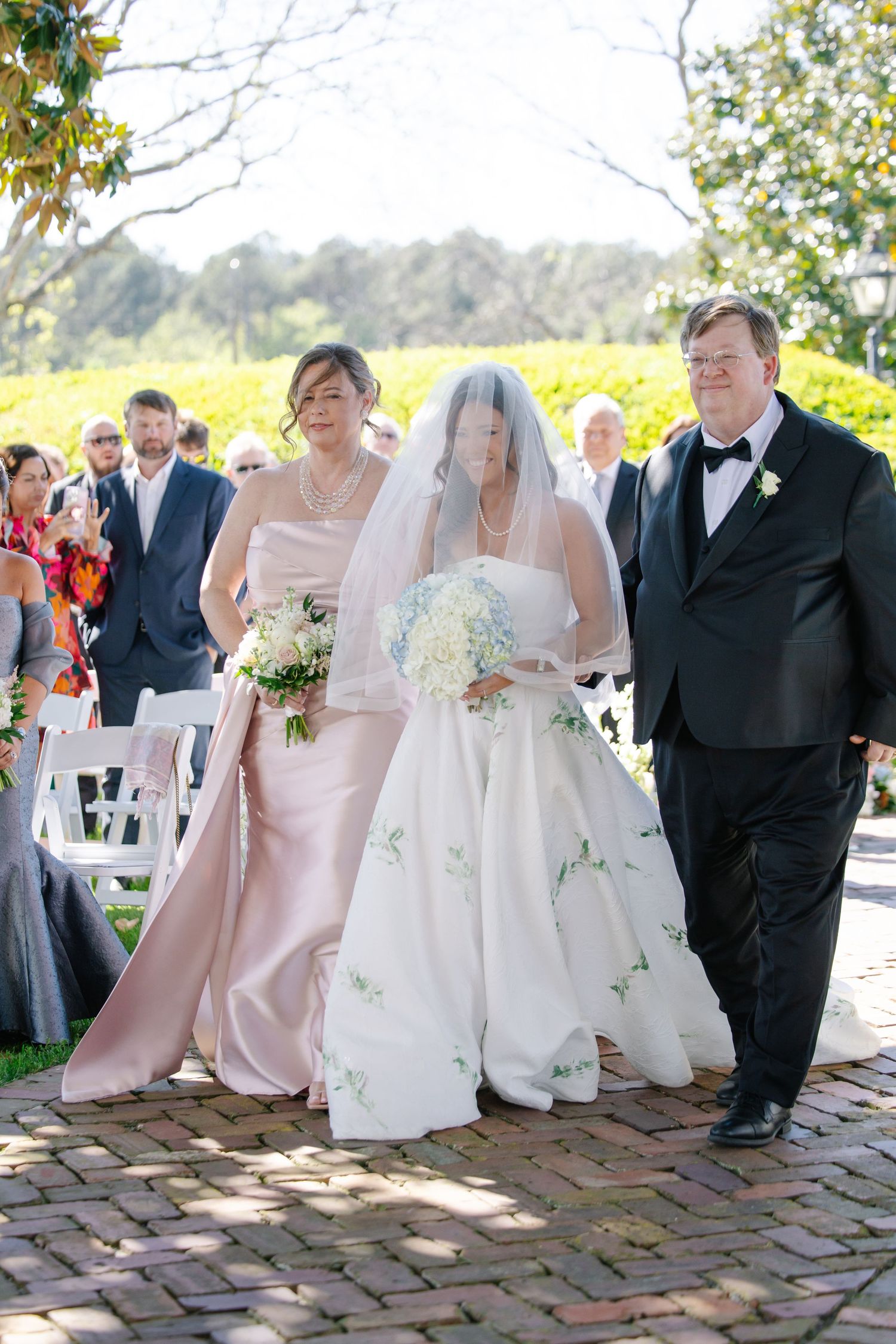 Bride in floral white gown walking down brick aisle flanked by parents at outdoor ceremony.