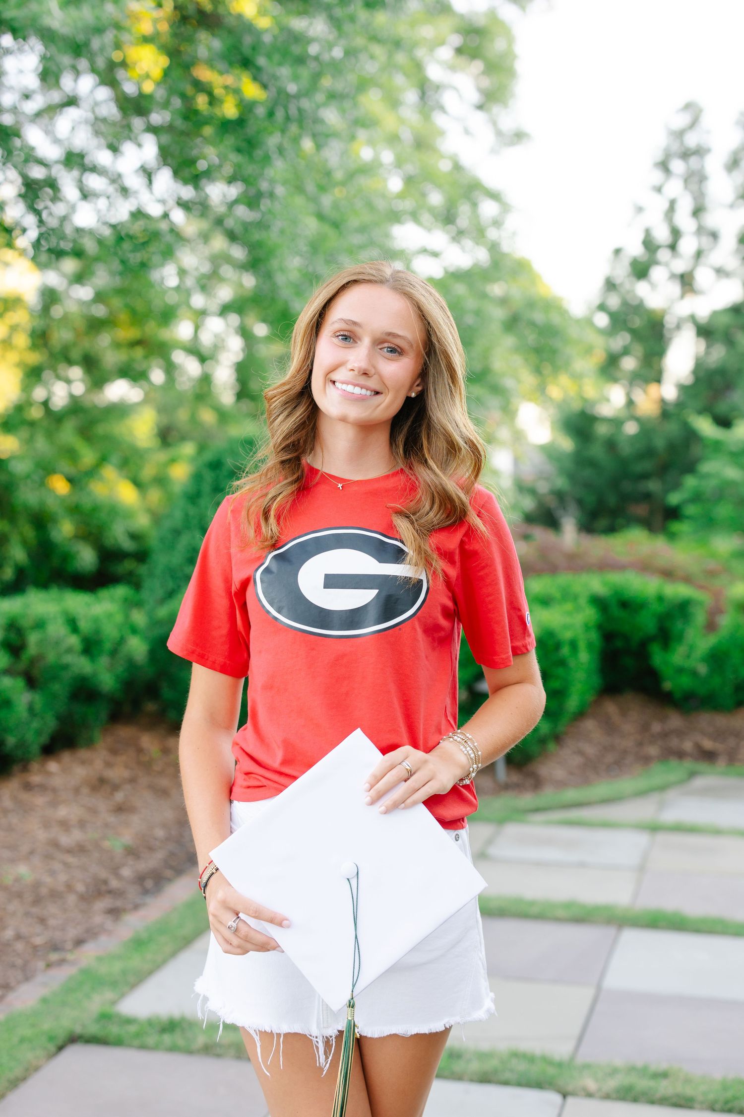 Smiling teen in red University of Georgia Bulldogs tee holding a diploma outdoors in a garden.