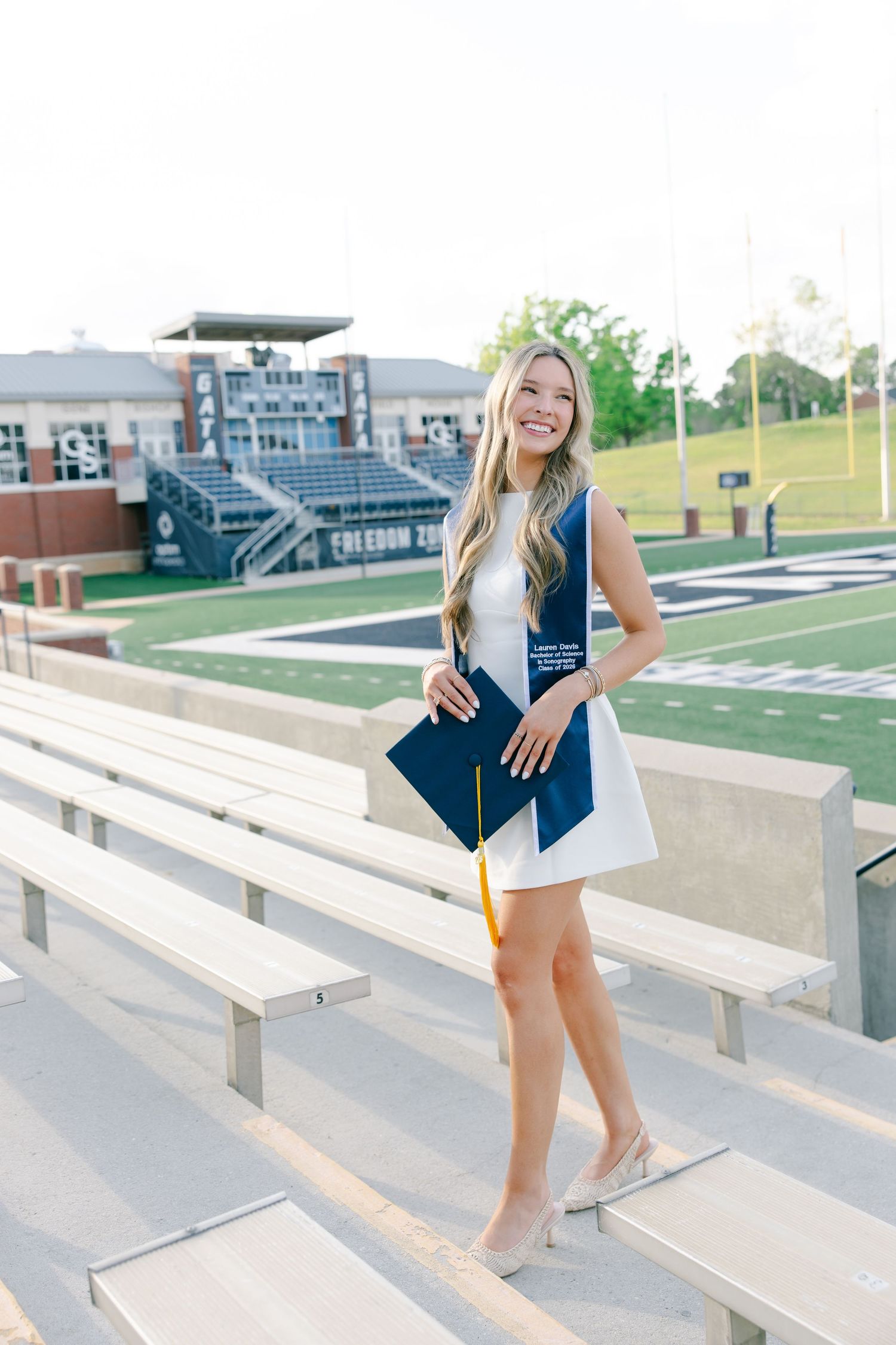 Happy female graduate in white dress holding navy cap with gold tassel at stadium bleachers.