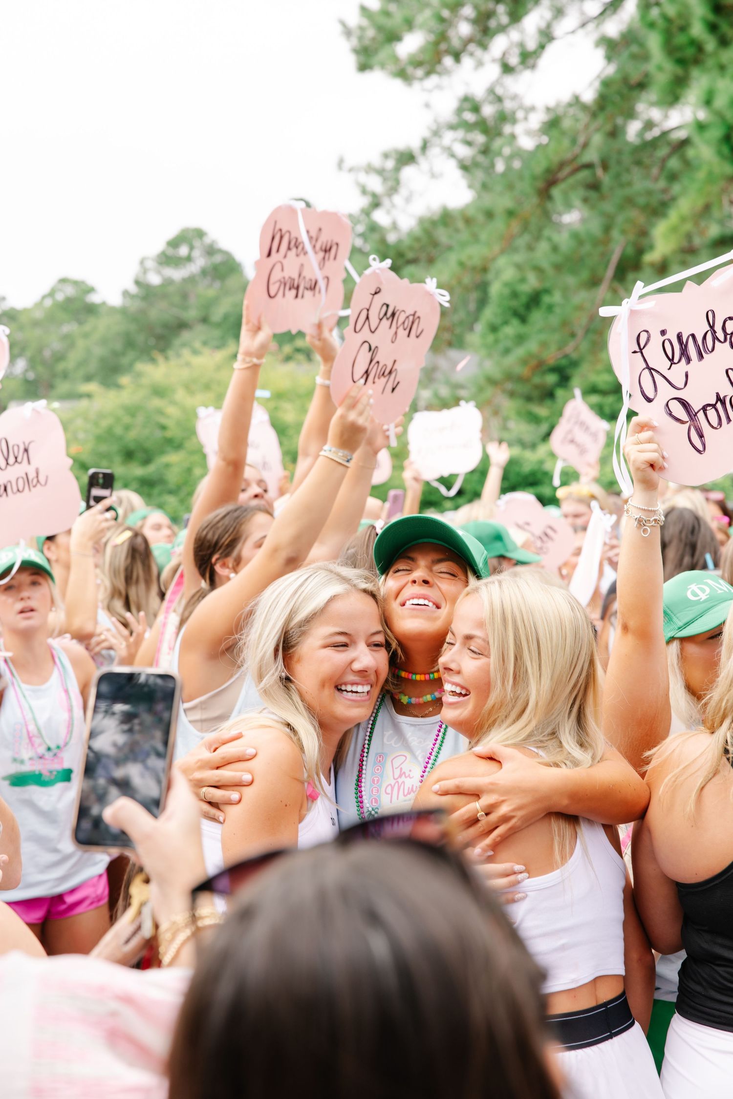 Excited sorority women embrace and celebrate during Phi Mu bid day with pink signs and confetti outdoors.