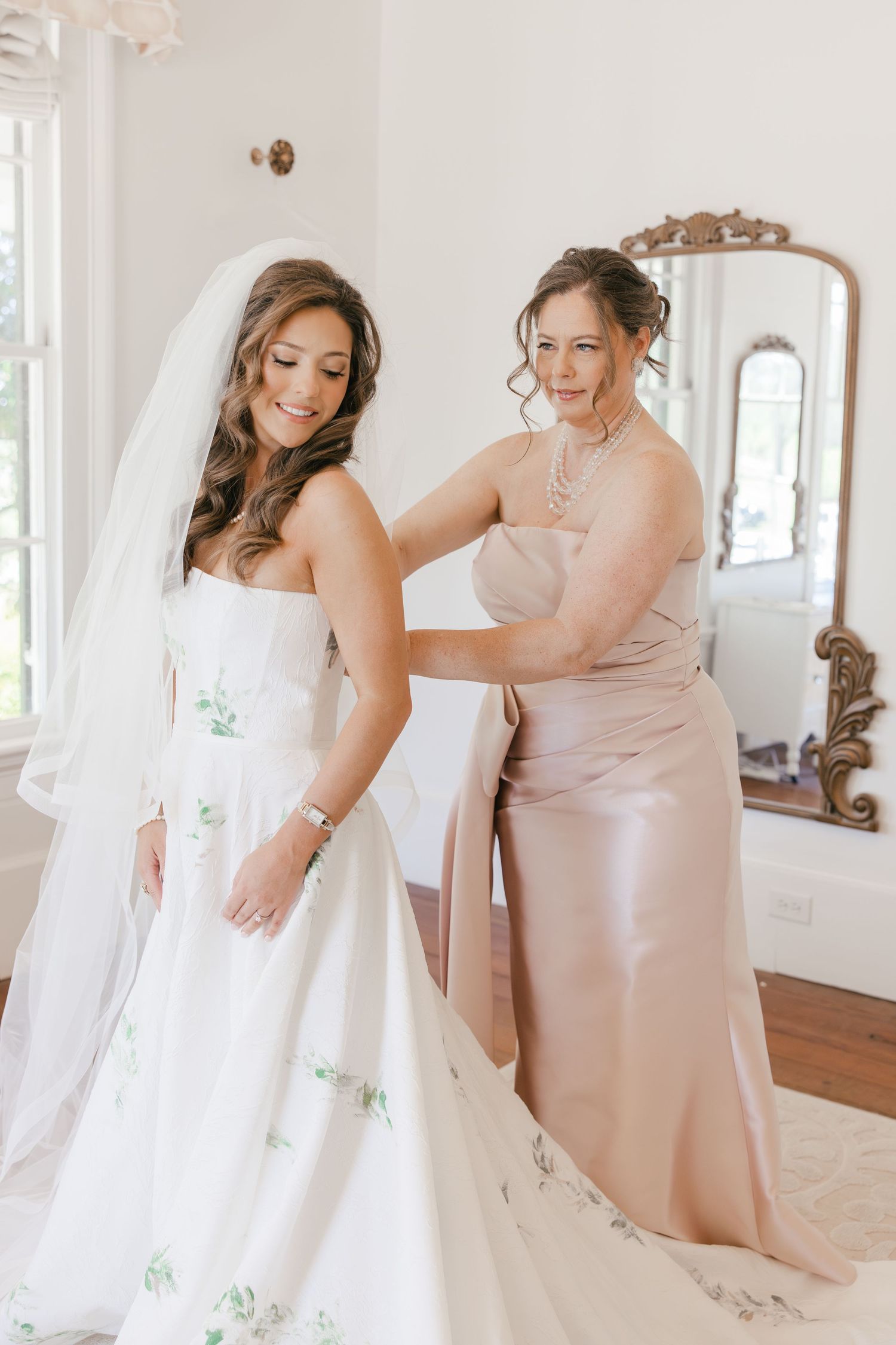 Mother helping bride into floral wedding gown with cathedral veil.
