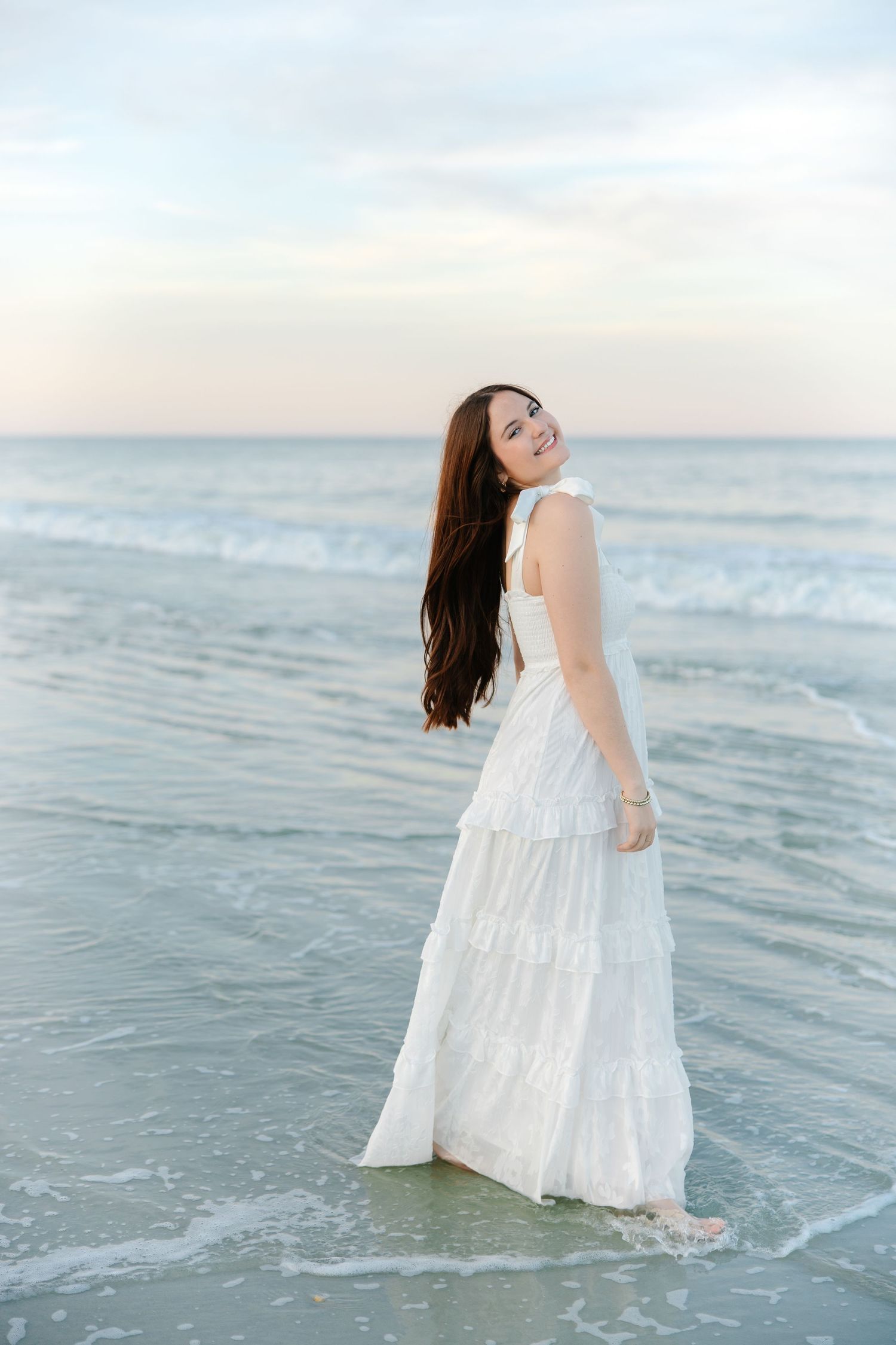 Joyful brunette woman in white maxi dress laughing while standing in gentle ocean waves at dusk.