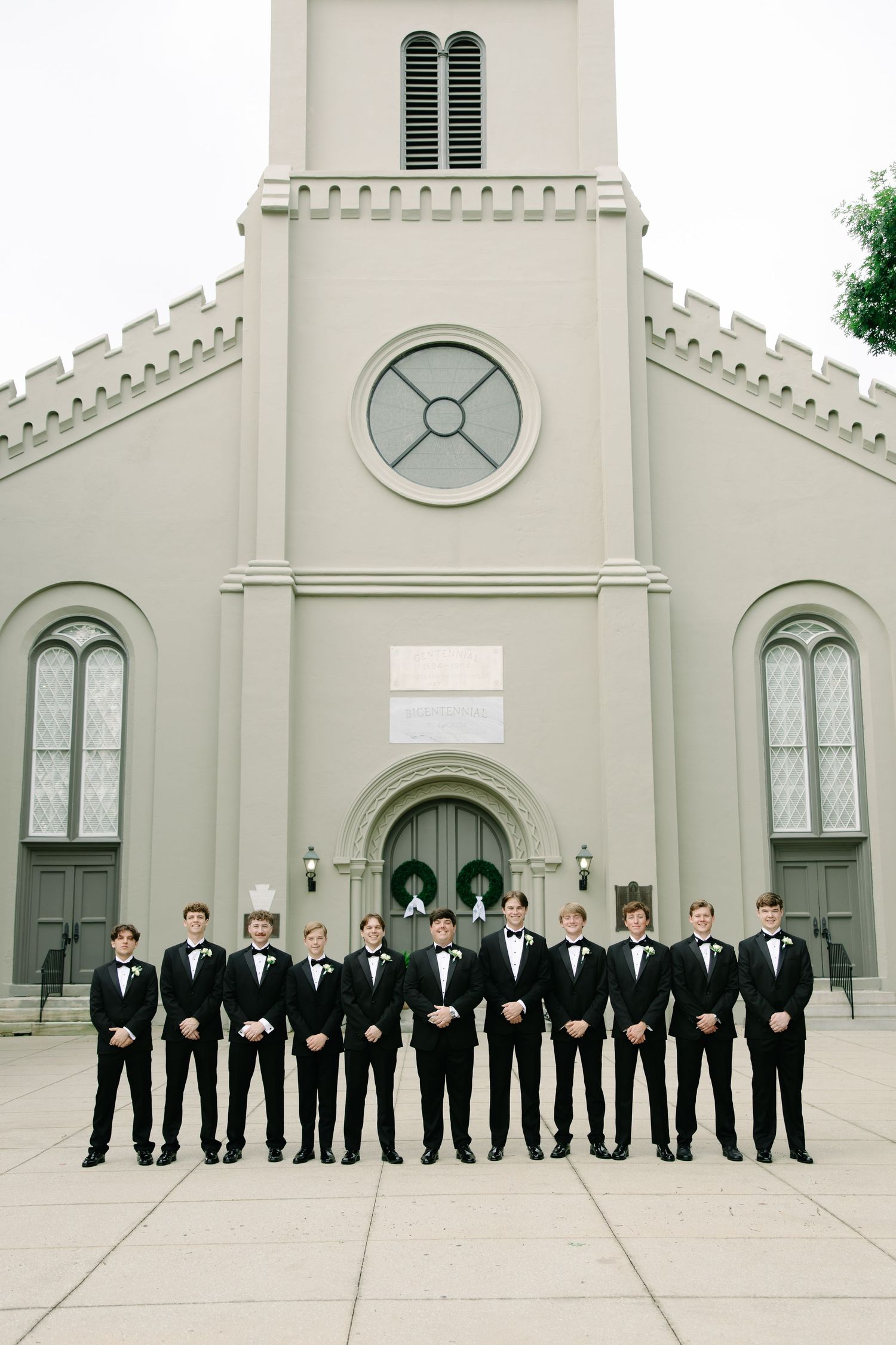 Groom and groomsmen in classic black tuxedos stand in formal line outside historic white church.