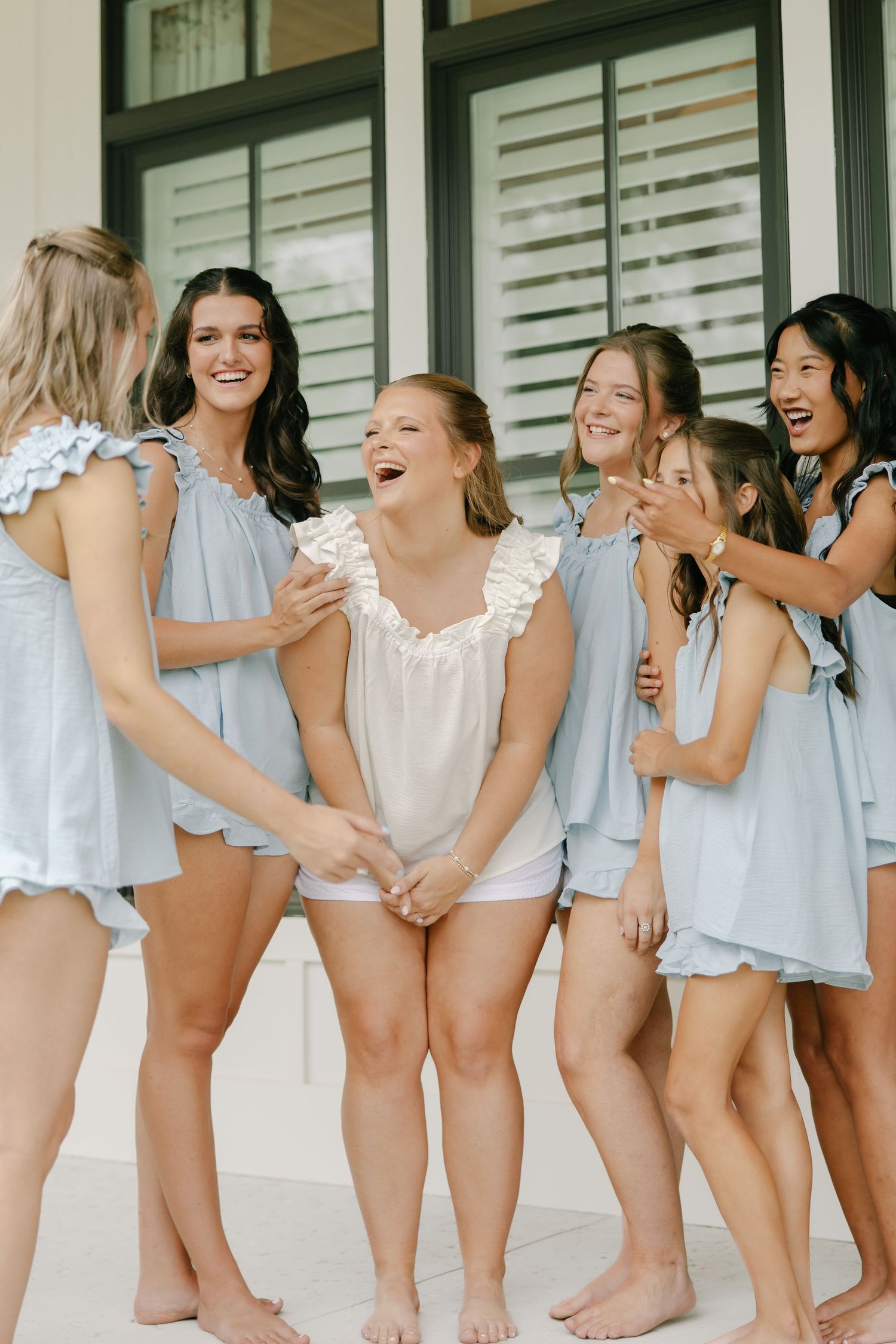 Bride laughing with bridesmaids in matching light blue ruffle rompers during wedding morning preparations.