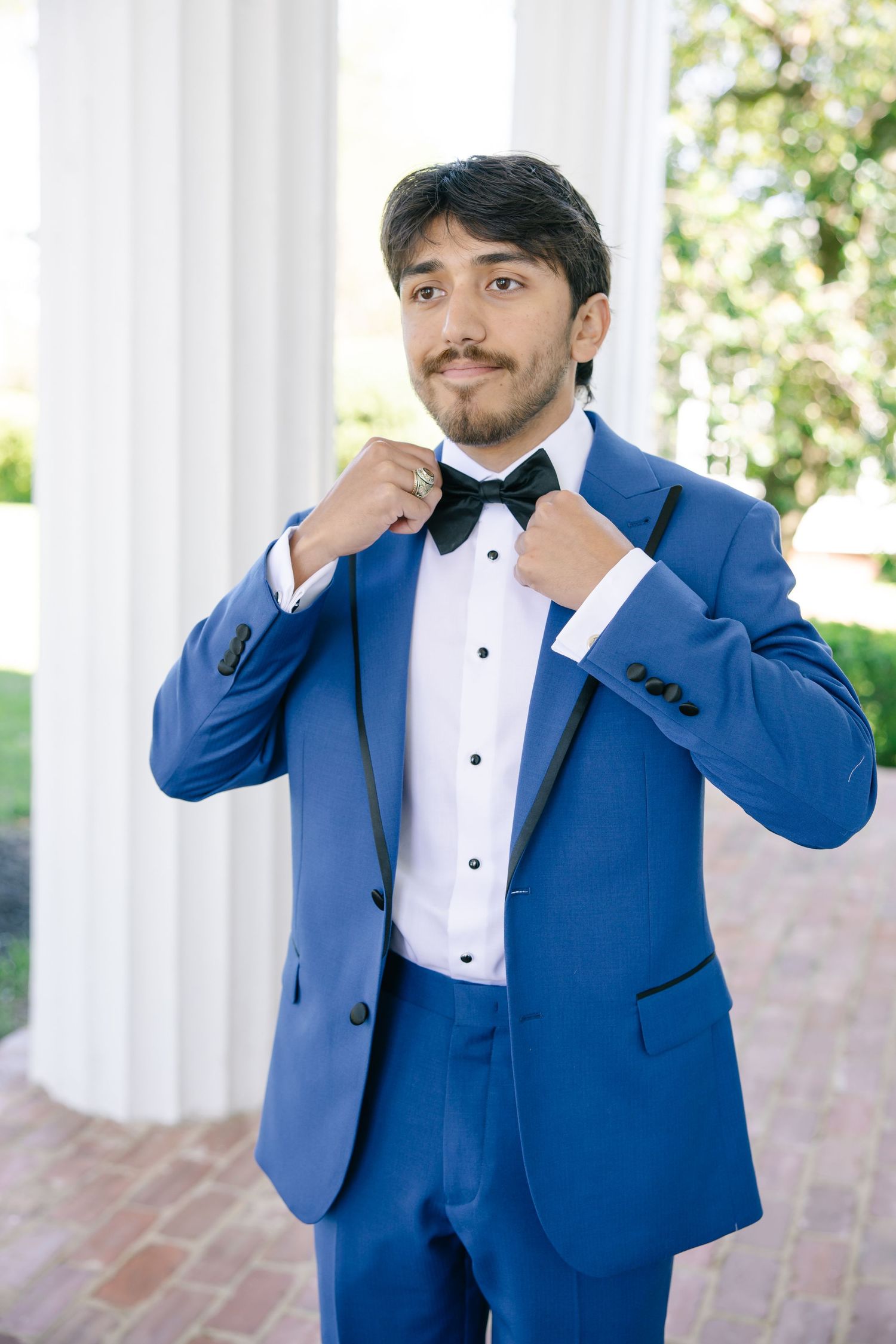 Dapper groom in royal blue tuxedo adjusting black bow tie between white columns outdoors.