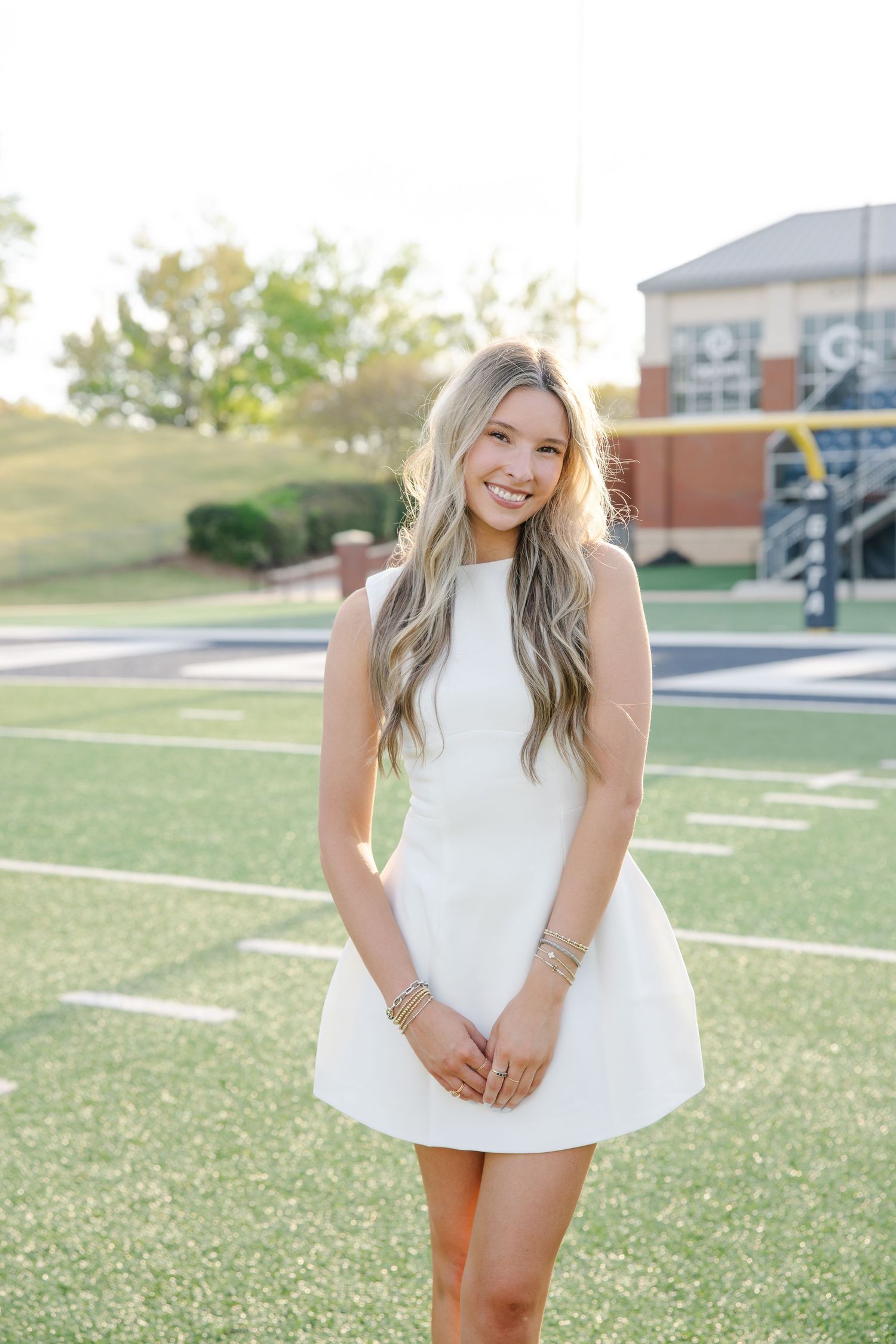 Blonde female graduate smiling in white mini dress standing on college football field at sunset.