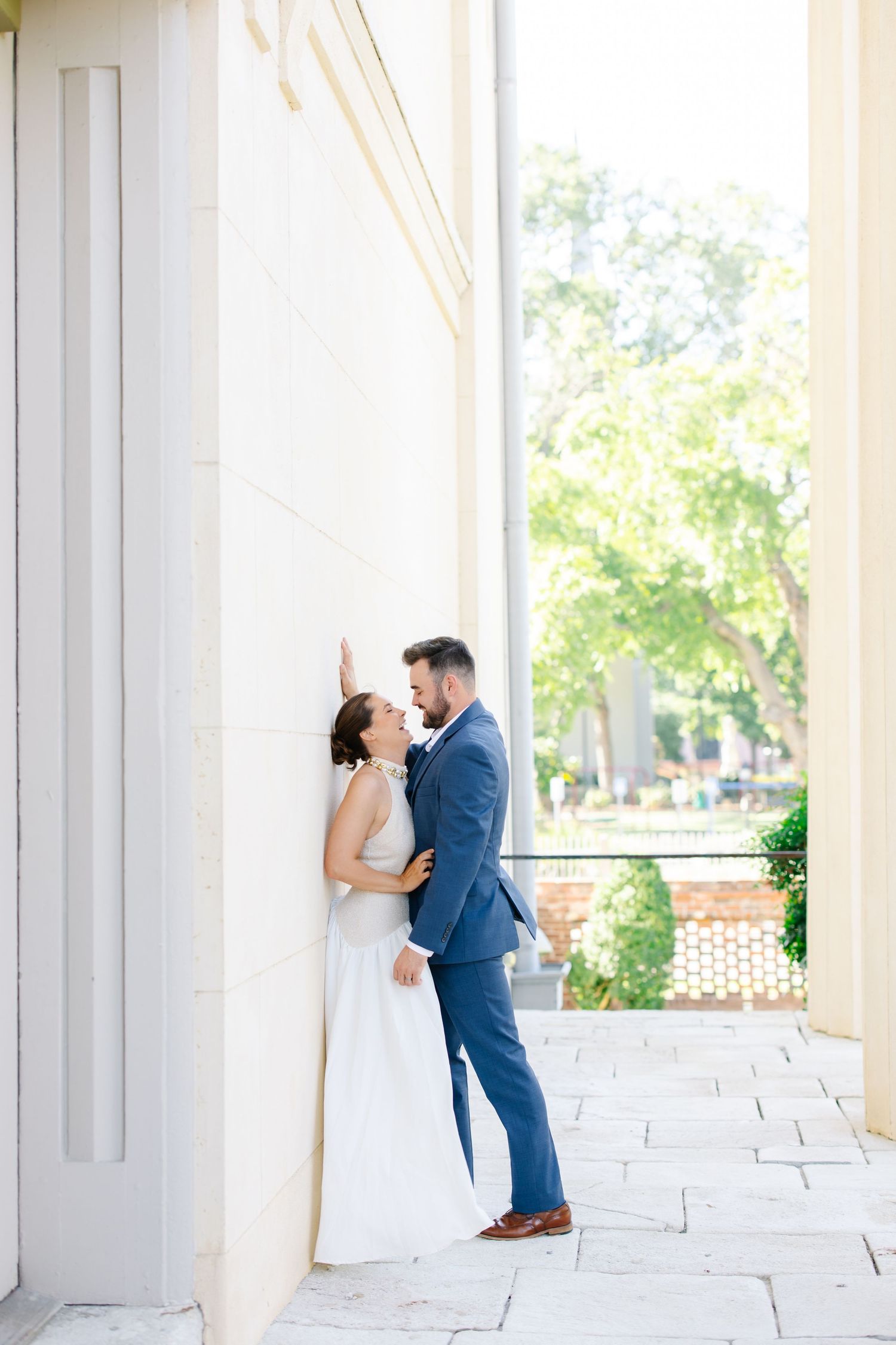 Bride and groom sharing romantic moment leaning against white column in sunlit outdoor corridor.