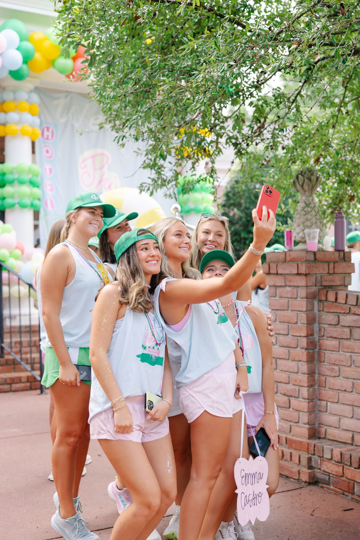 Group of sorority women in matching green caps take a selfie at a colorful bid day celebration outdoors.