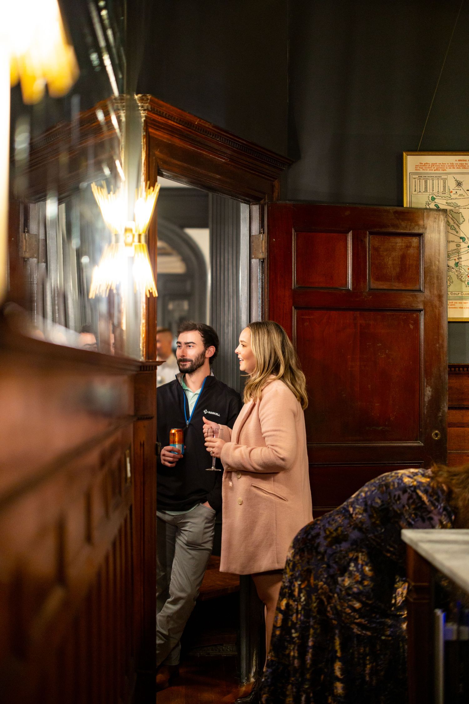 Couple holding champagne glasses exploring a warmly lit historic bar with rich dark wood paneling.