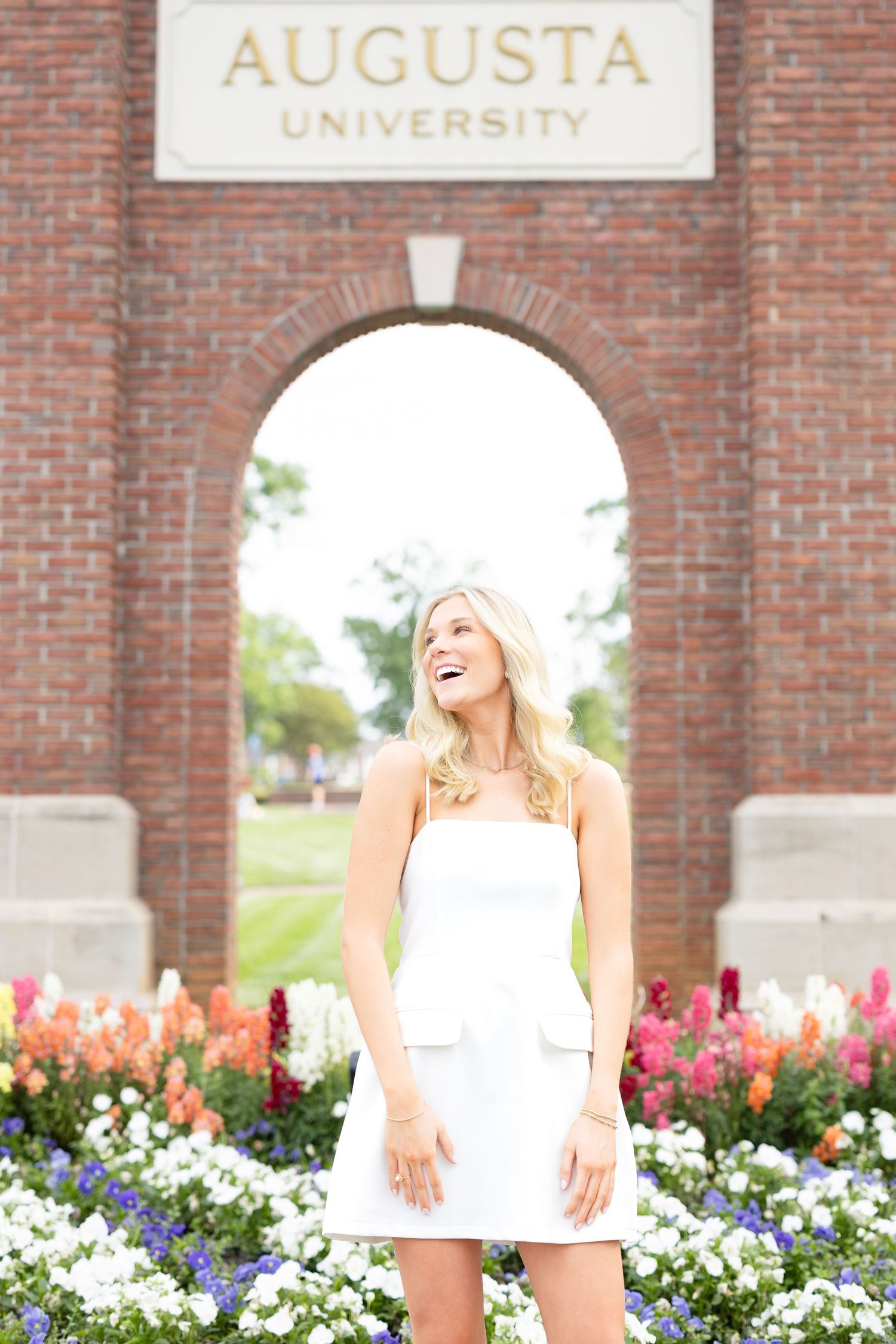 Blonde graduate in white dress laughing joyfully under Augusta University arch with vibrant flowers.