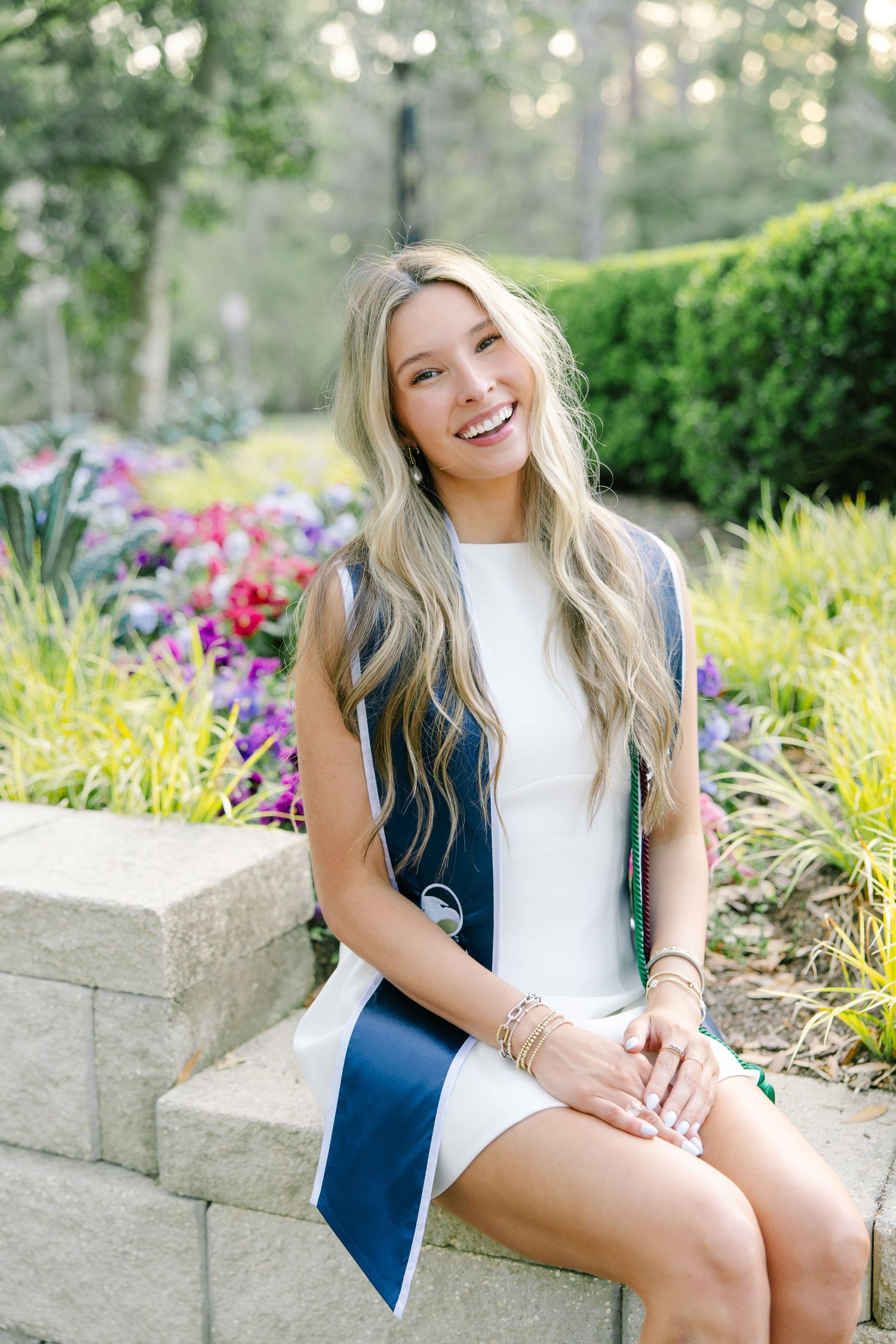 Happy college graduate in white dress with navy graduation stole sitting on stone wall beside colorful flowers.