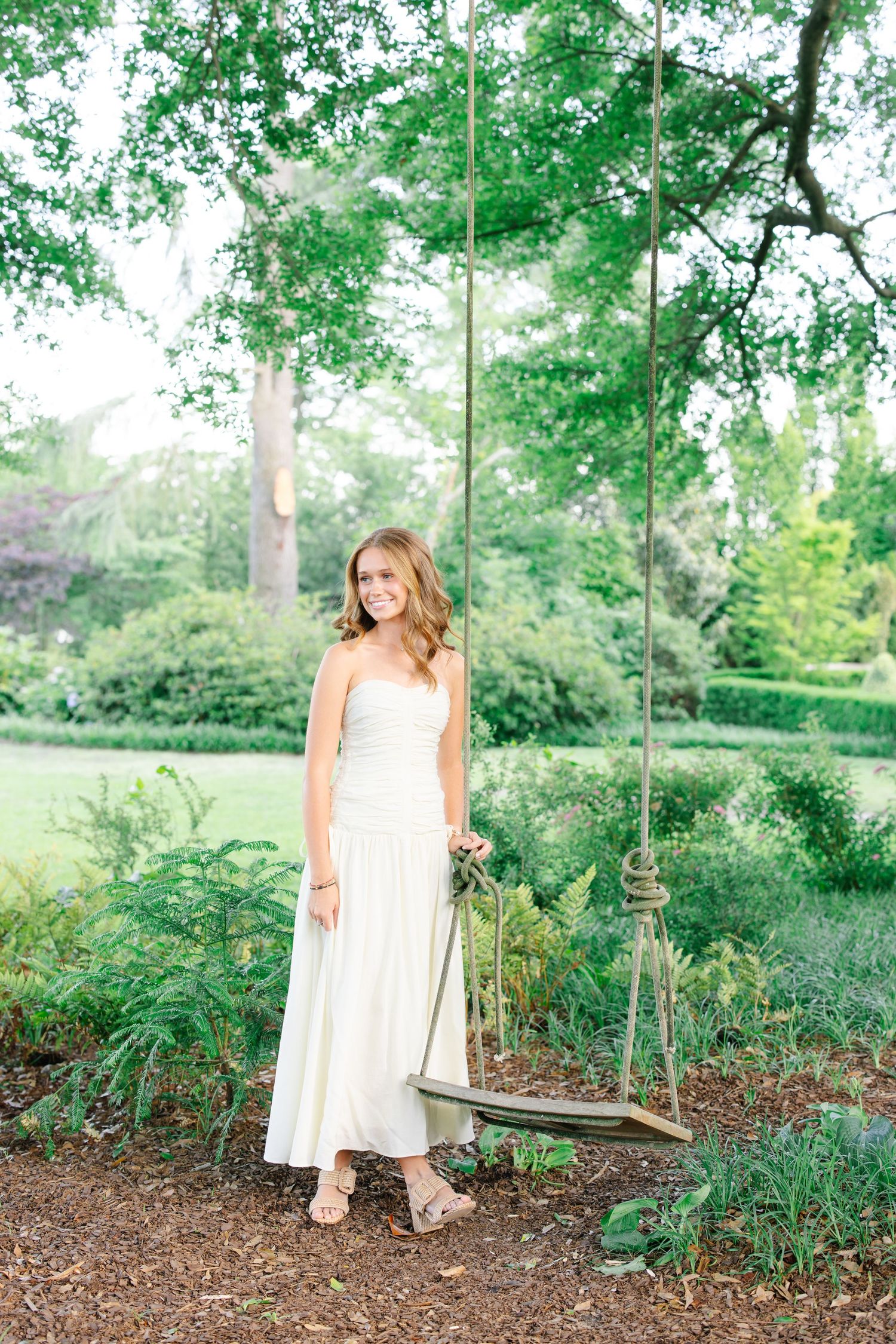 Young woman in white strapless maxi dress standing beside a rope tree swing in a lush garden.