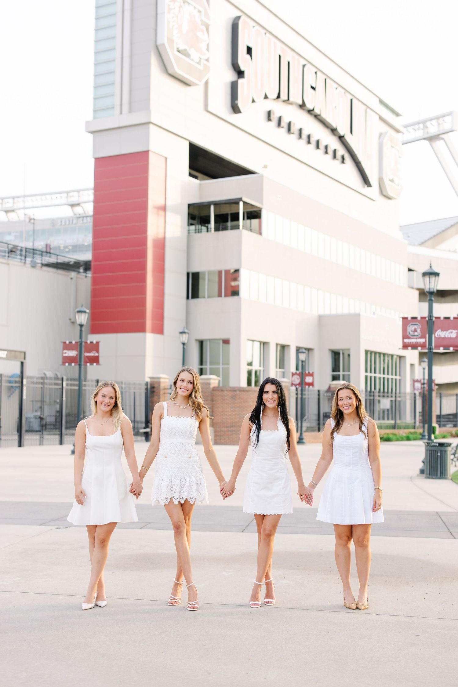 Four young women in white dresses holding hands walking in front of a stadium entrance.