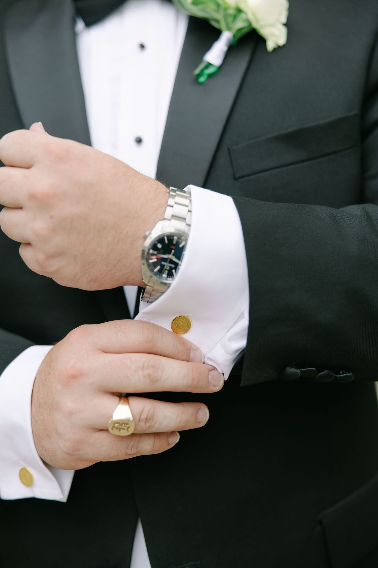 Groom in black tuxedo adjusting cufflinks wearing gold signet ring and silver watch on wedding day.