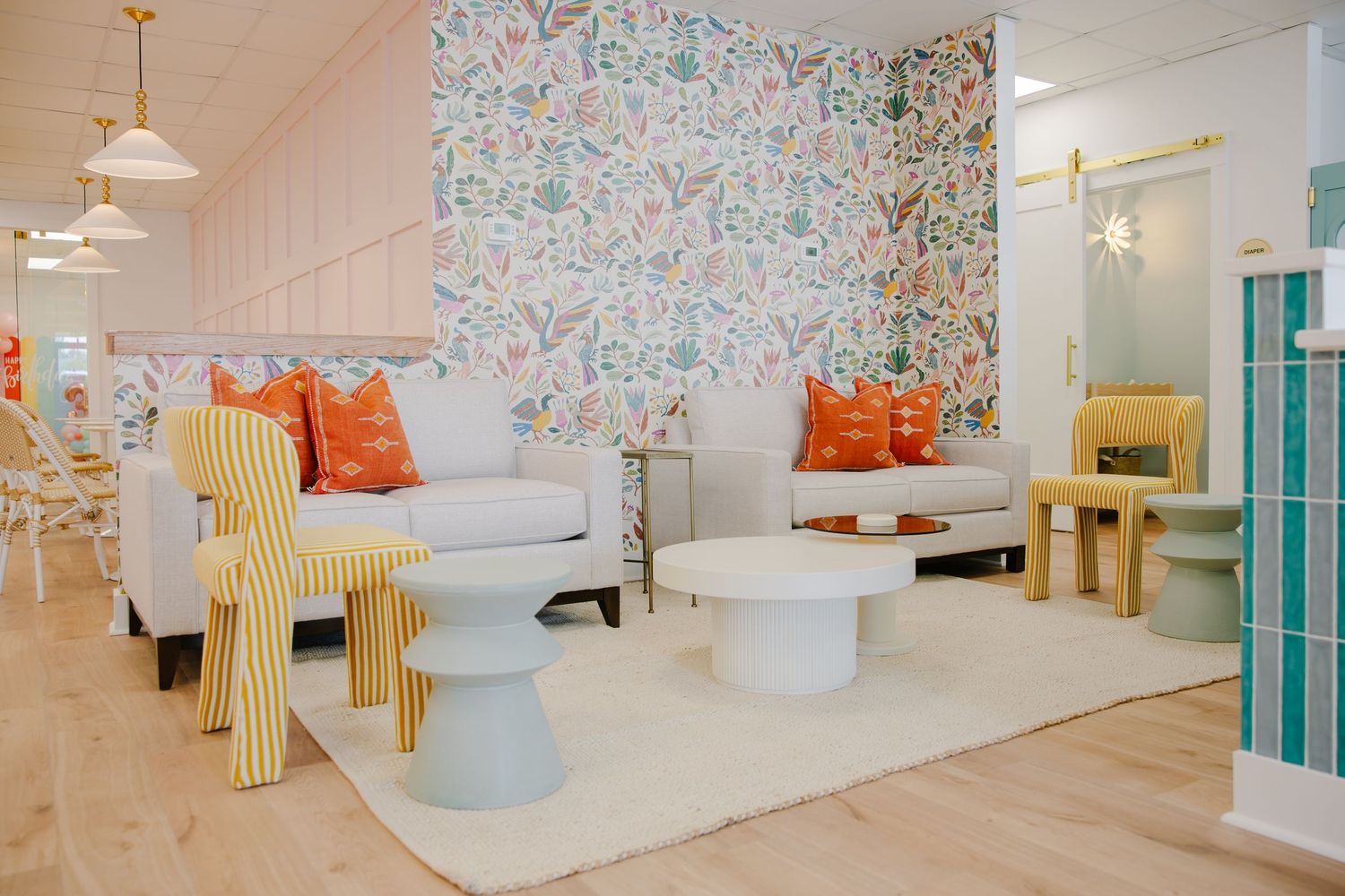 Bright playroom lounge with floral wallpaper, white sofas, orange pillows, and colorful modern furniture.