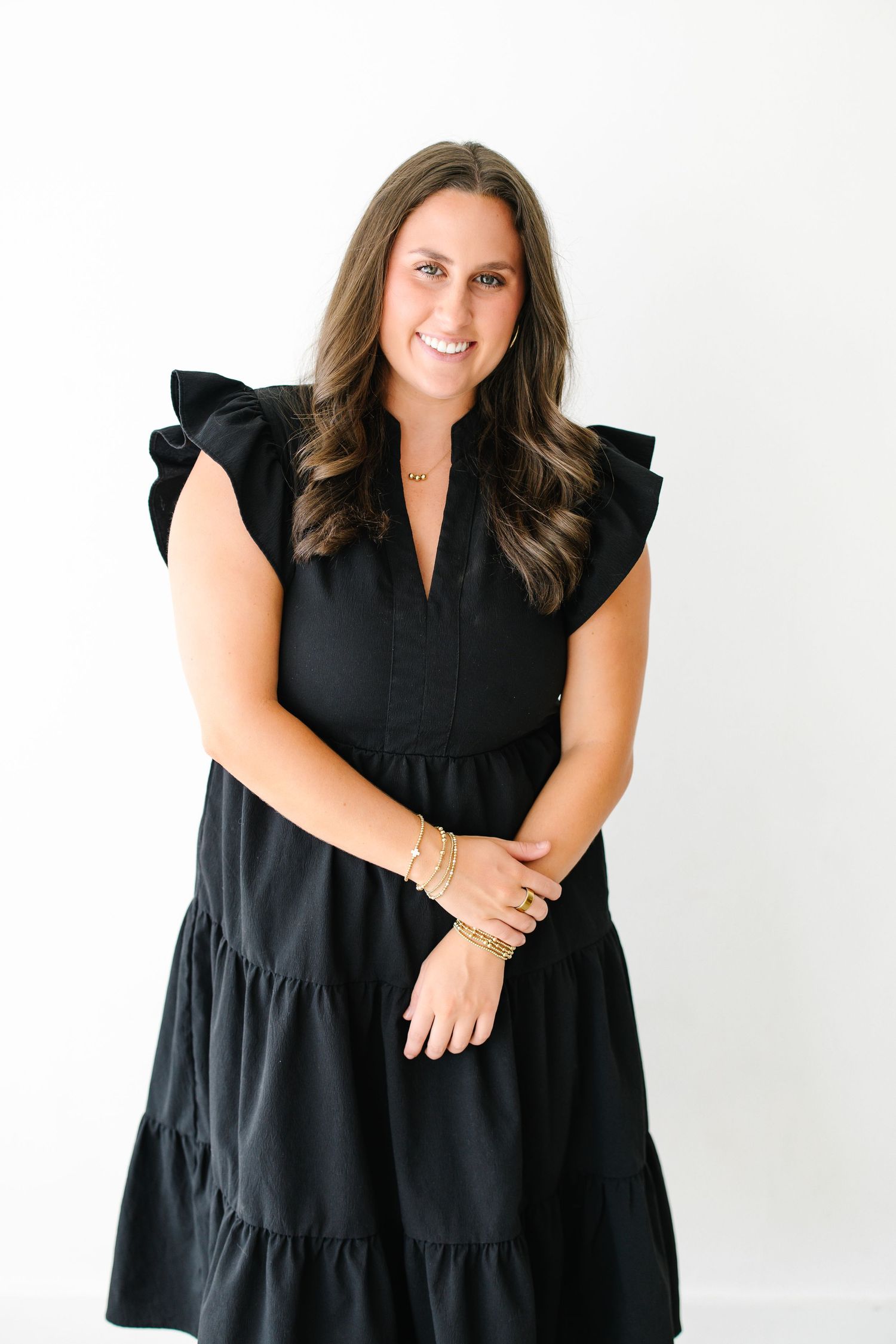 Smiling brunette woman in black ruffled dress with gold jewelry poses against white background.