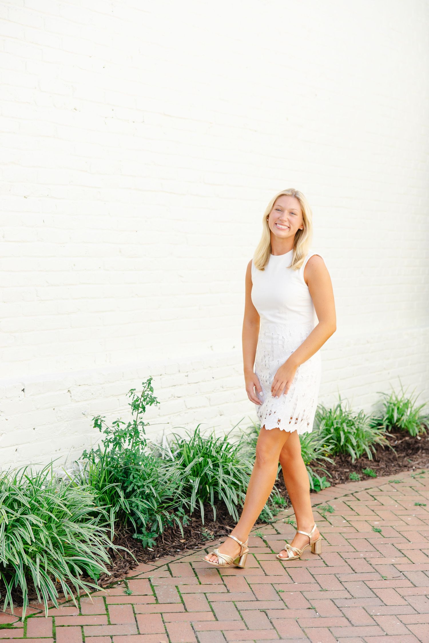 Smiling blonde woman in white lace mini dress walking along brick path by white wall.