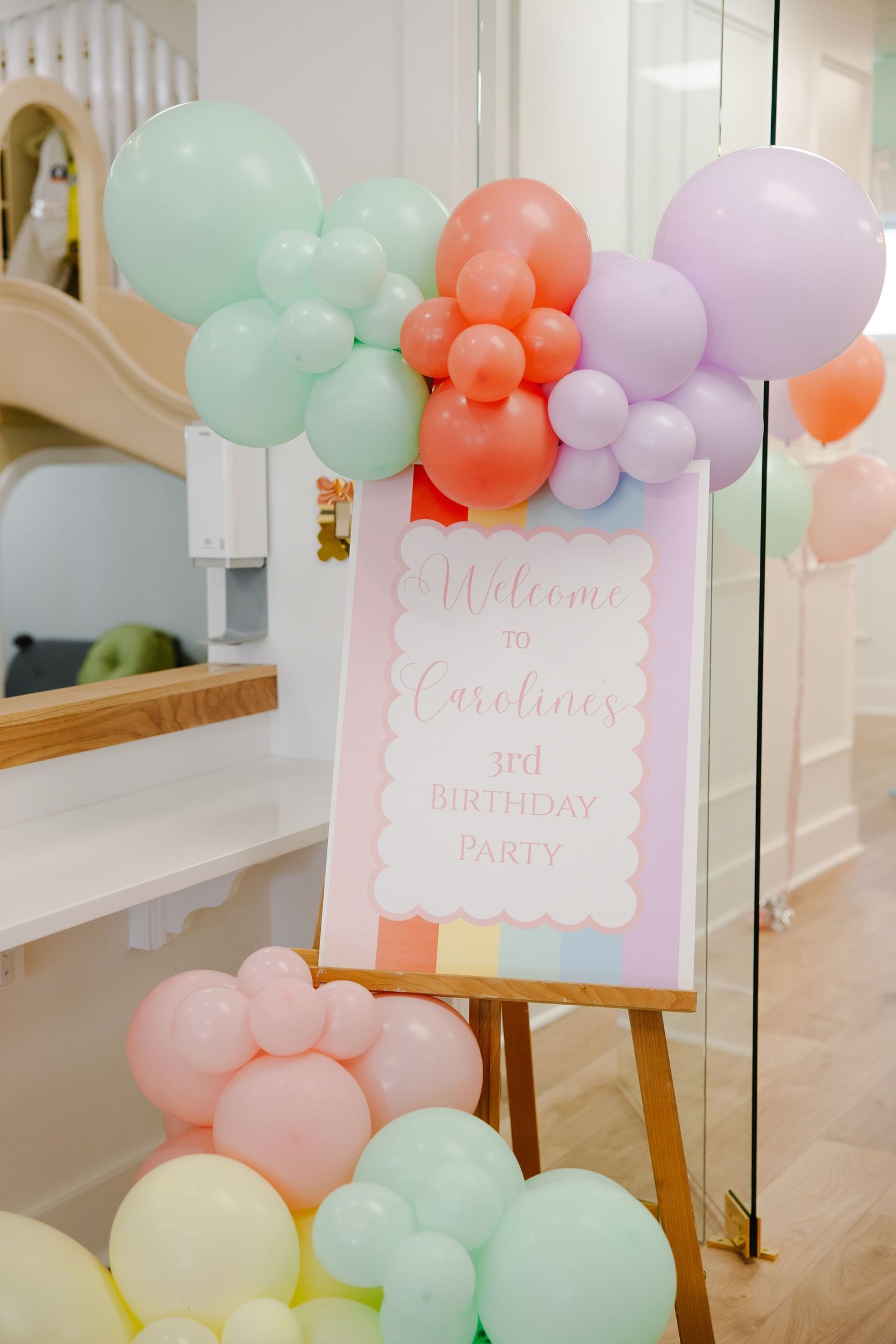 Welcome sign for Caroline's 3rd birthday party surrounded by a colorful pastel balloon garland display.