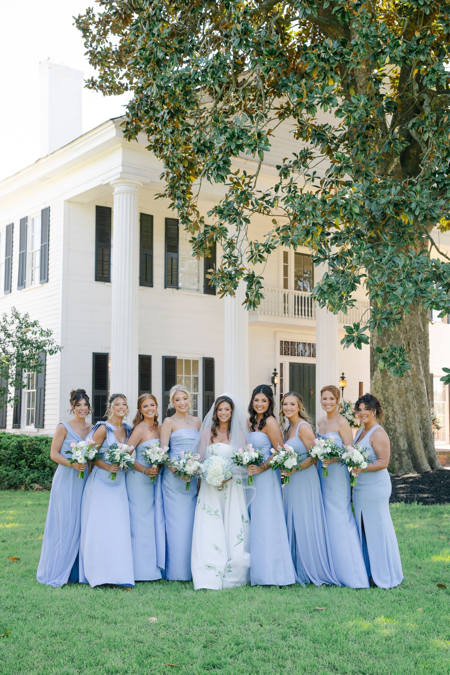 Bride in floral white gown poses with eight bridesmaids in periwinkle blue dresses holding white bouquets outside elegant estate.