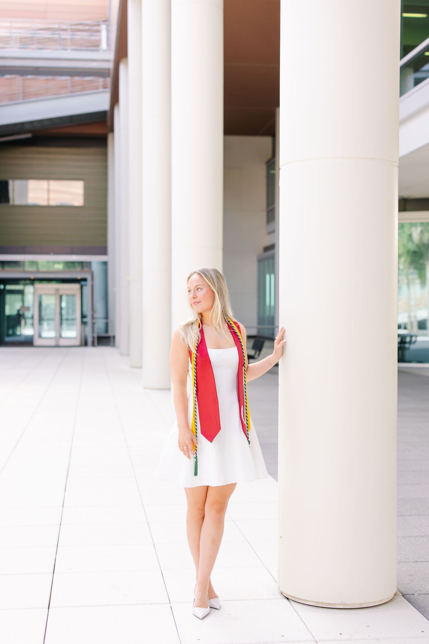 Blonde graduate in white dress with honor stoles leaning against column at university.