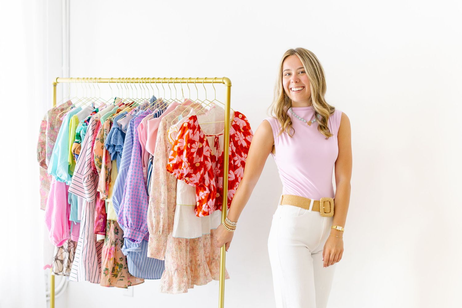 Smiling blonde woman in pink top and white jeans standing beside gold rolling rack of colorful clothes.