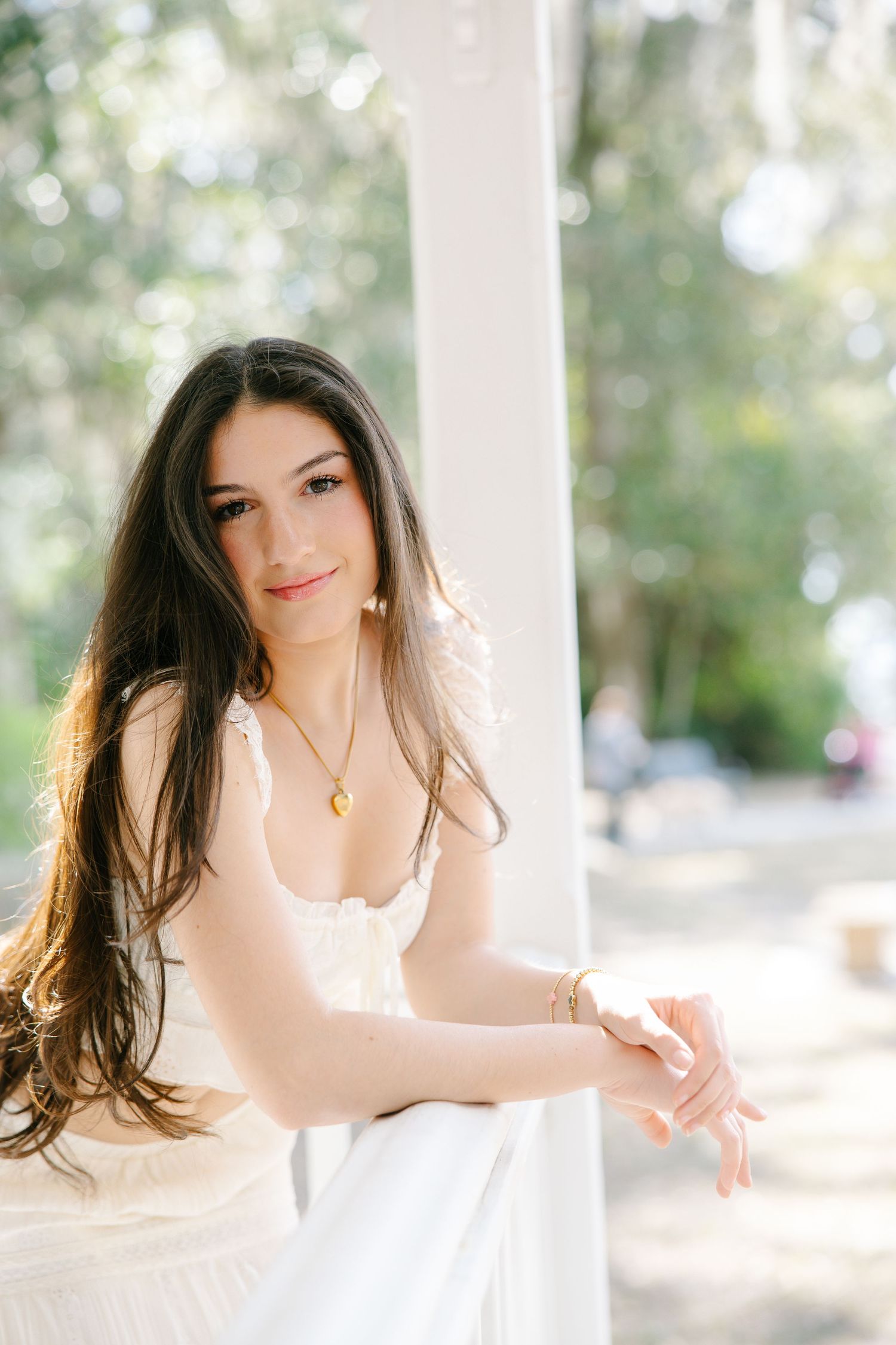 Brunette woman in white dress leaning against white column on a bright porch.