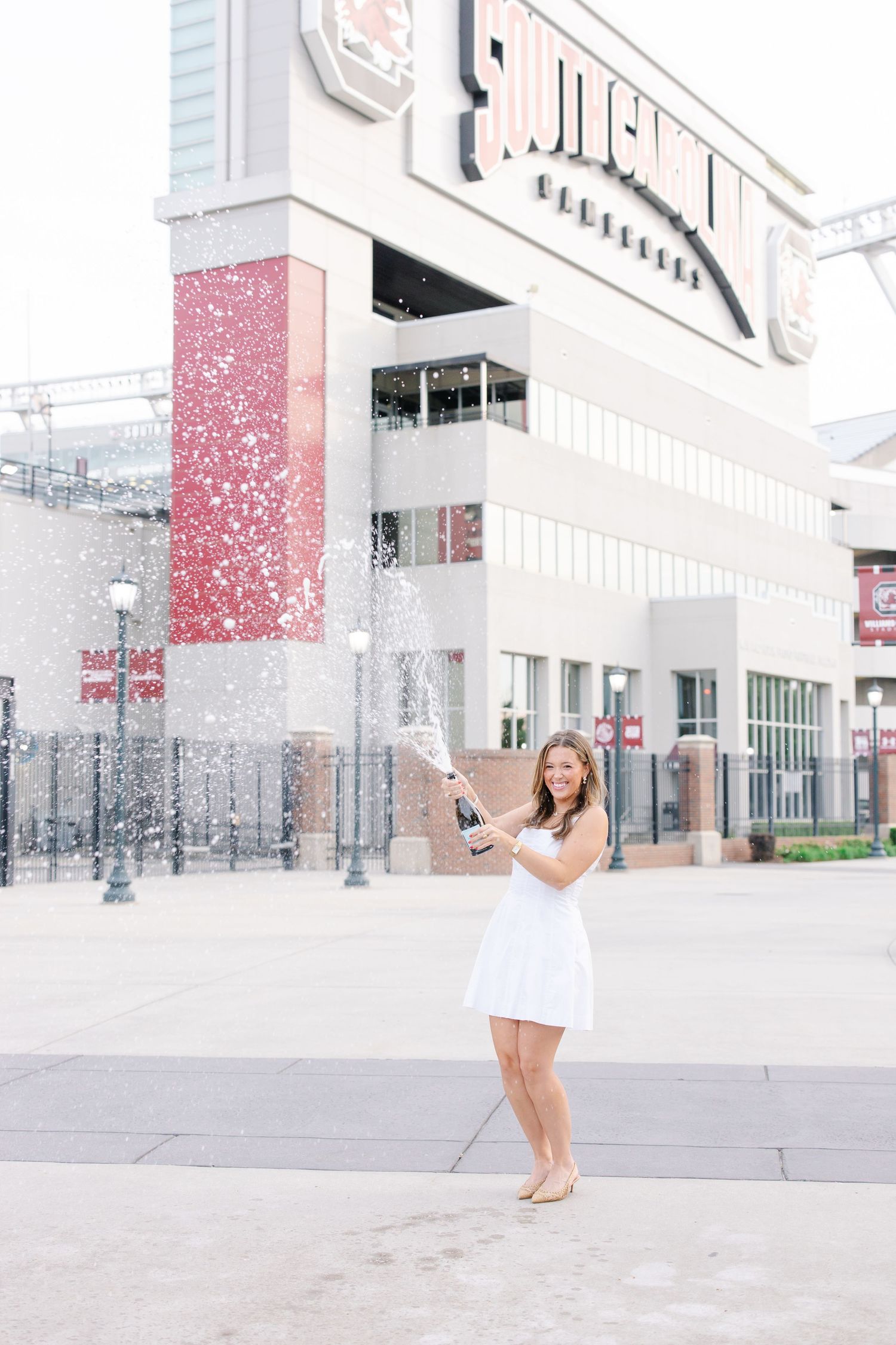 Woman in white dress popping champagne bottle outside South Carolina Gamecocks stadium.