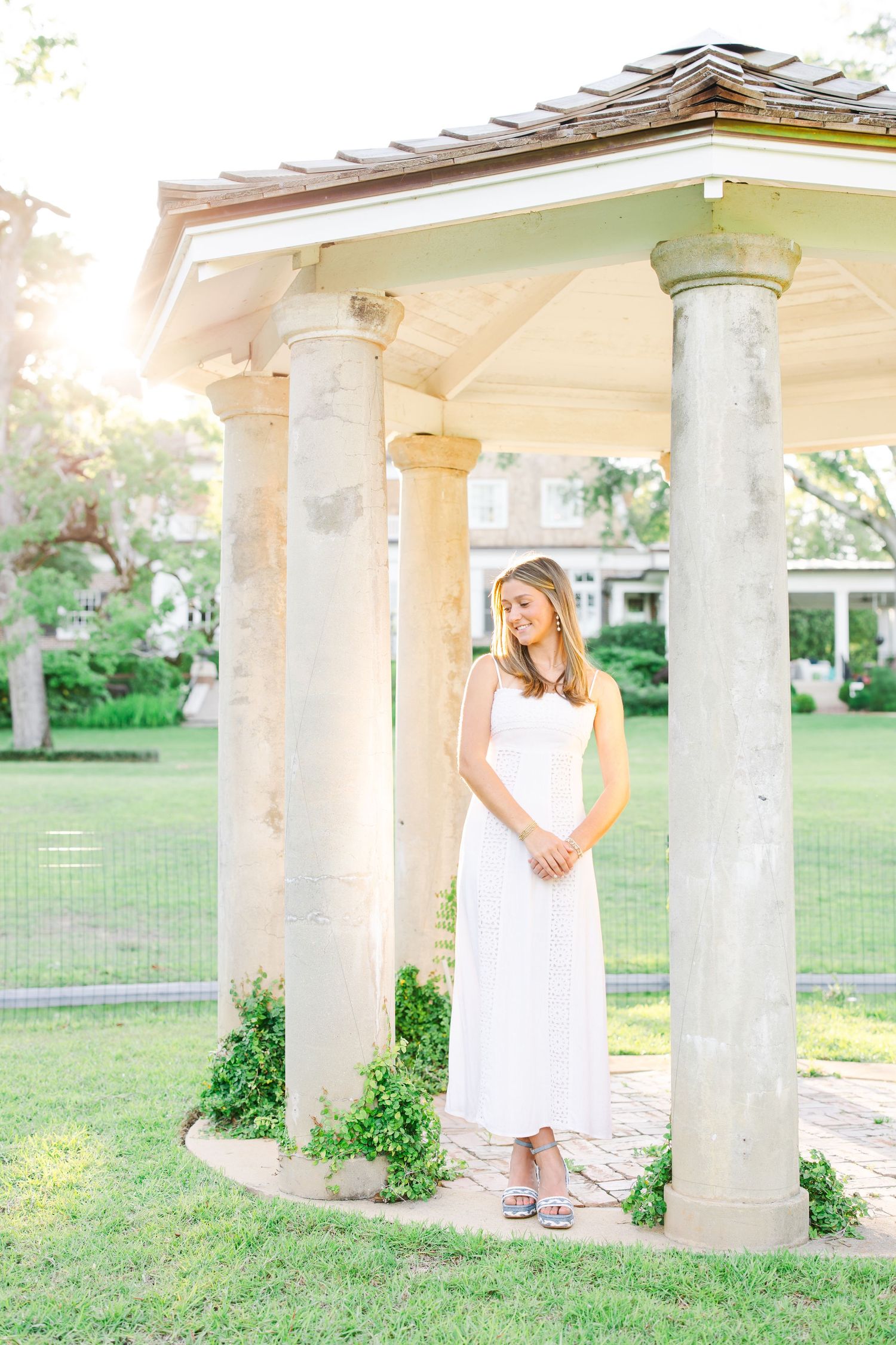 Young woman in white maxi dress standing between stone columns of a garden gazebo in soft sunlight.