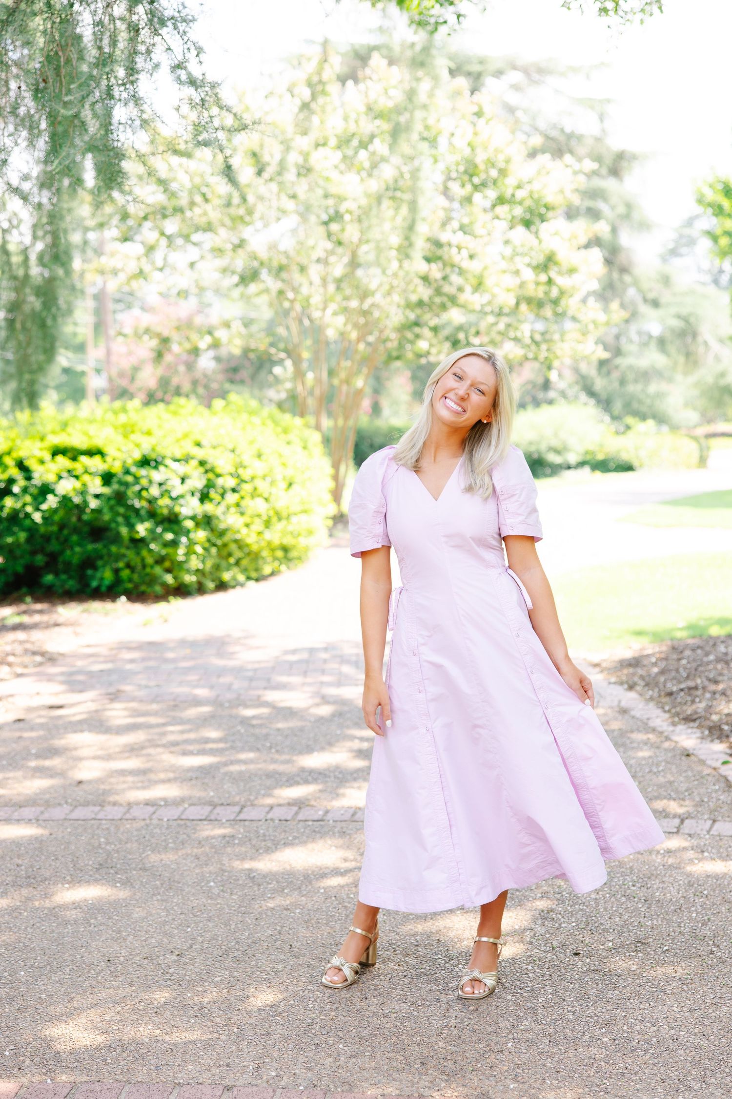 Blonde woman in pink midi dress twirling on garden path surrounded by green trees.