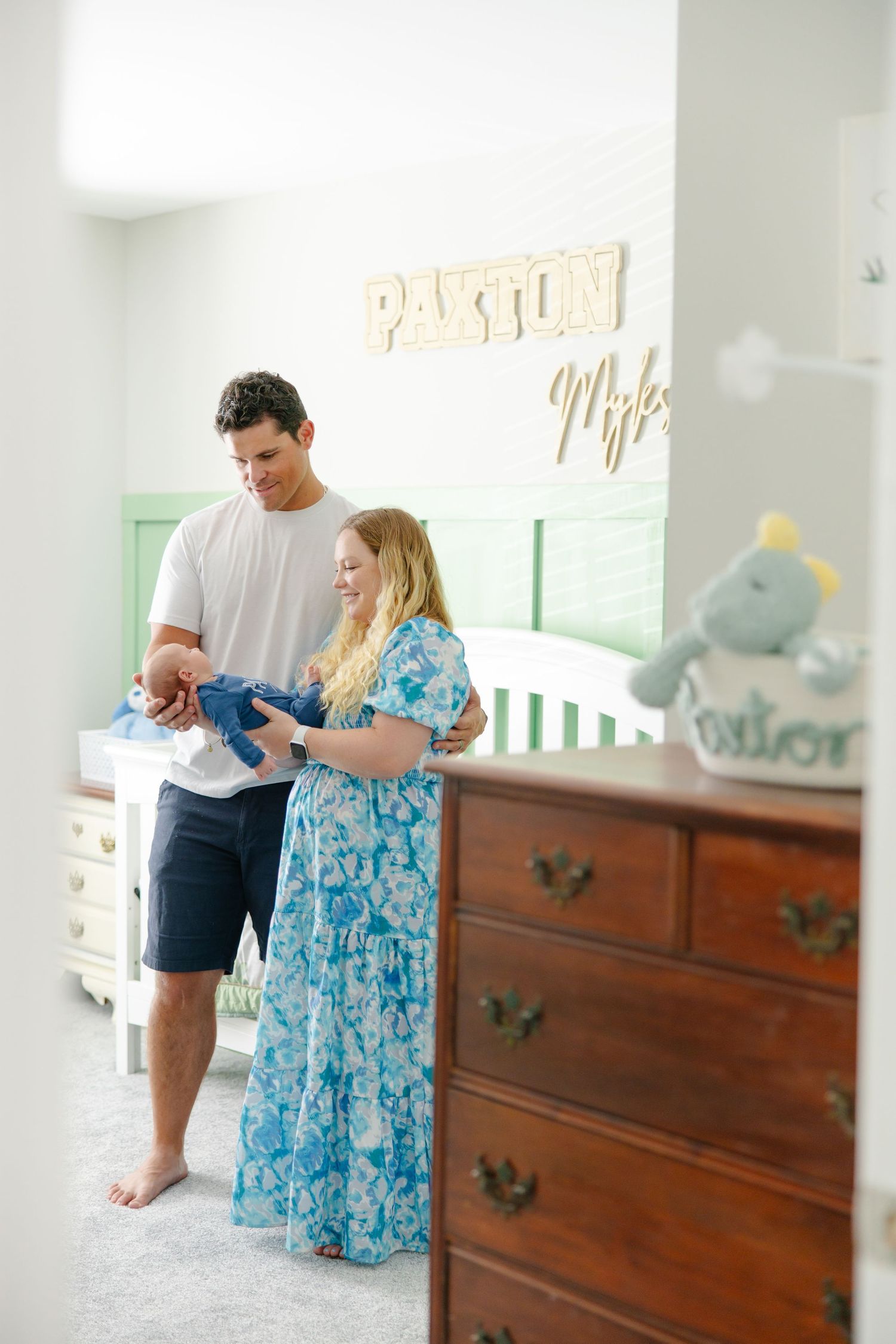 Couple standing in nursery holding baby clothes near crib with Paxton Miles name sign on wall.