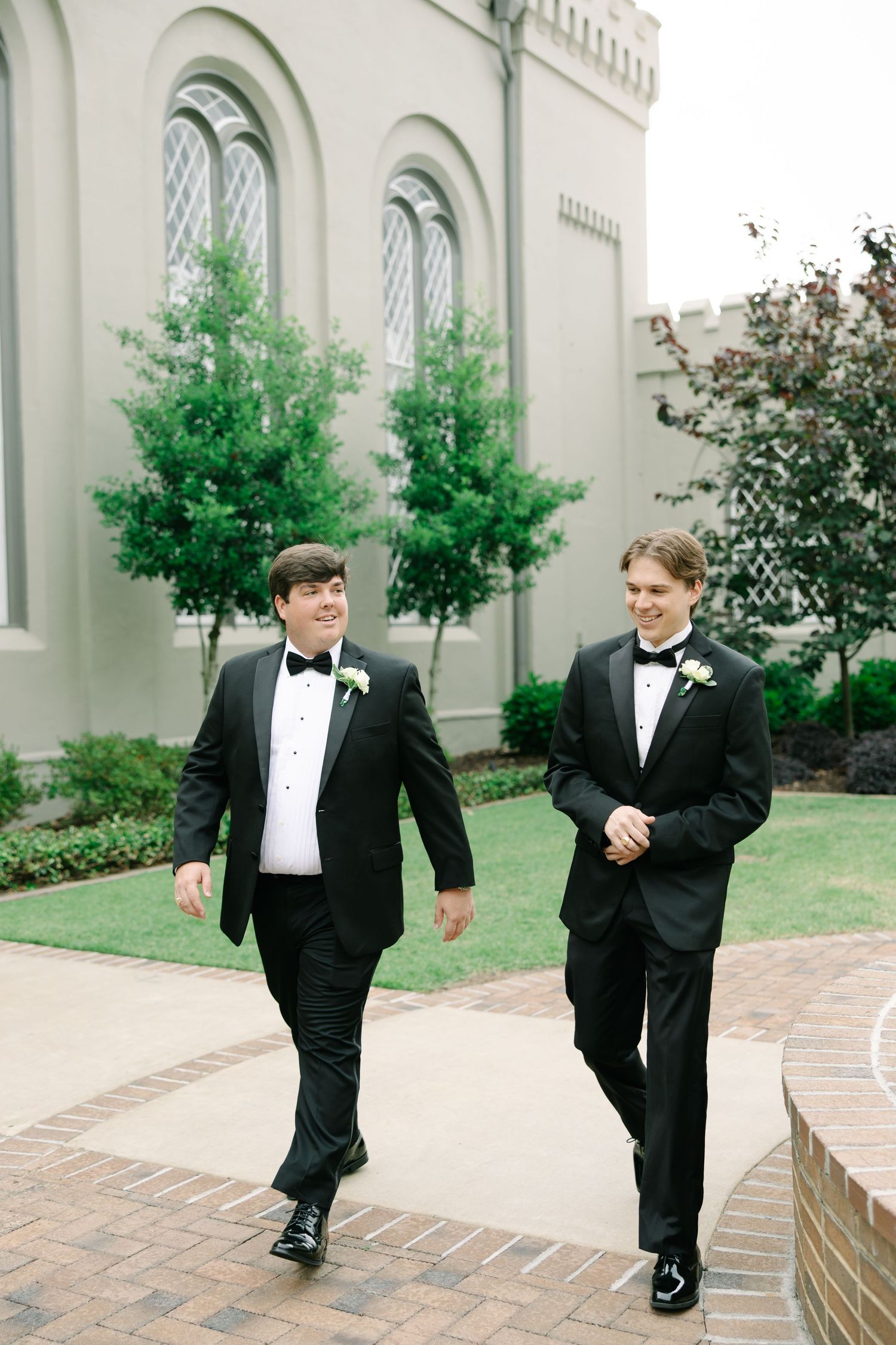 Two groomsmen in black tuxedos with boutonnieres walk and laugh outside a elegant stone church venue.