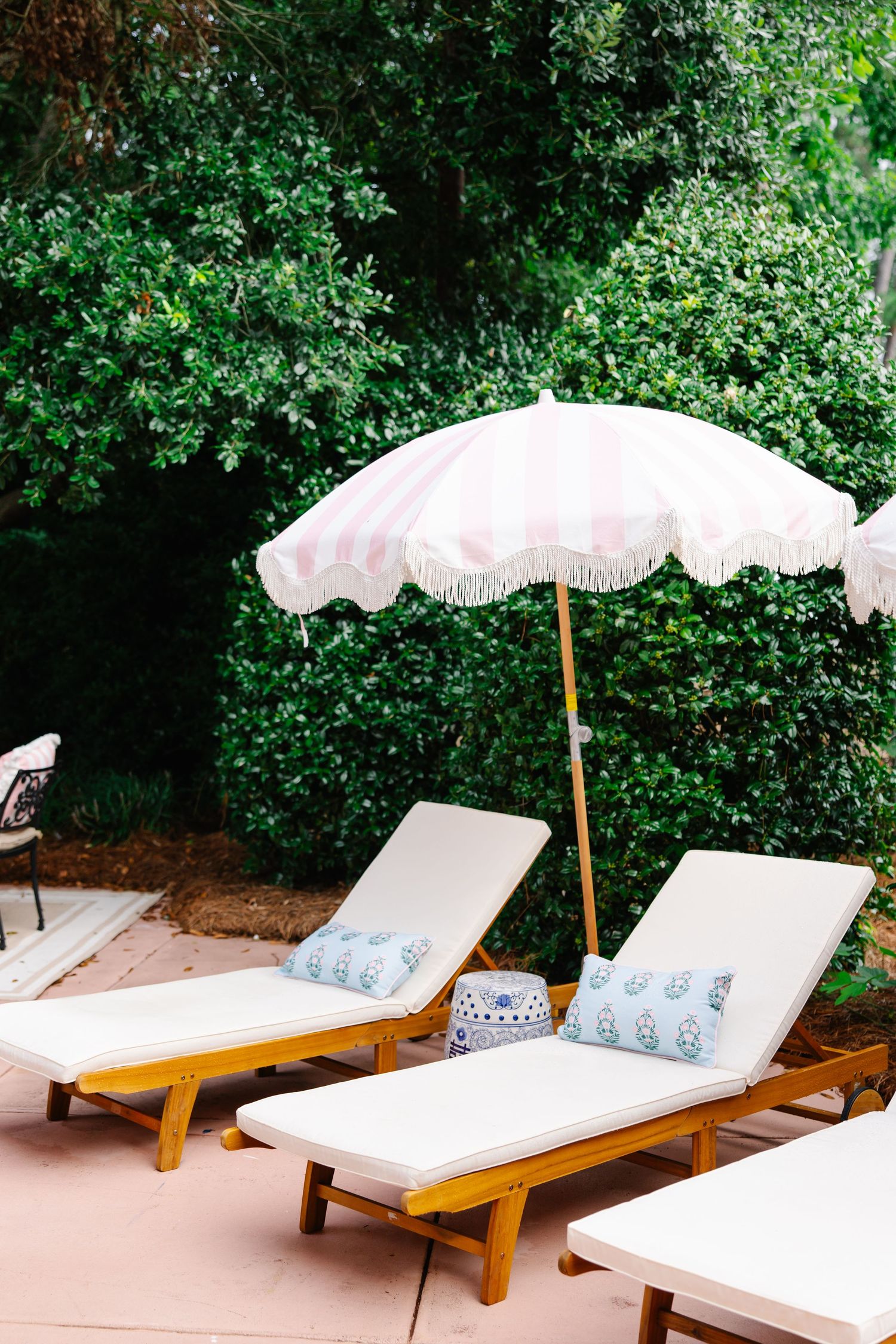 Two white cushioned lounge chairs with blue pillows under a pink fringed umbrella against lush green hedges.