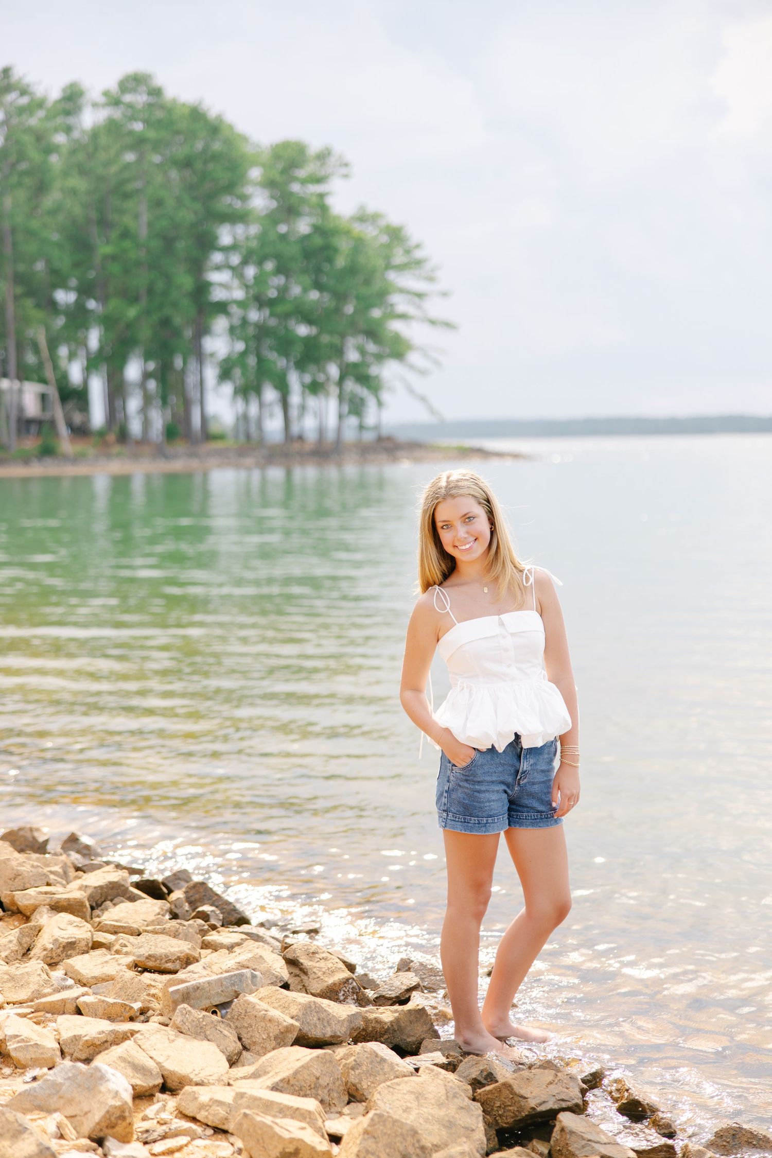 Smiling blonde teen in white ruffled top and denim shorts standing barefoot on rocky lake shoreline.