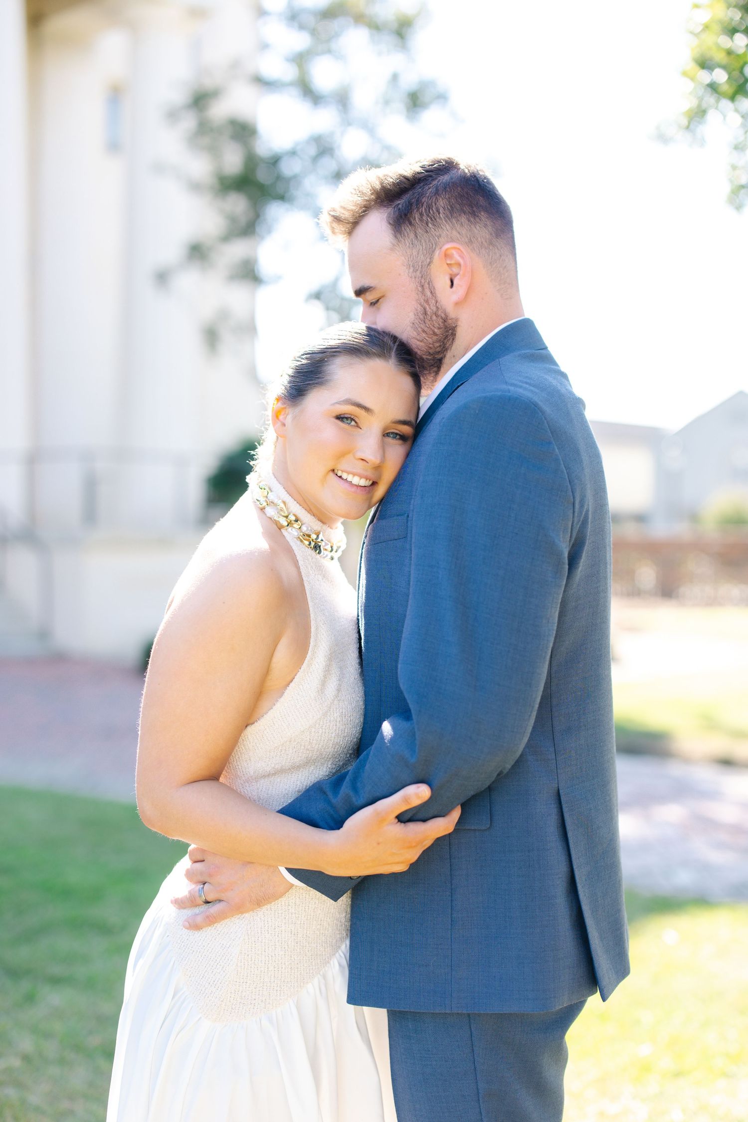 A groom in blue suit embraces a smiling bride in white halter gown outdoors in bright sunlight.