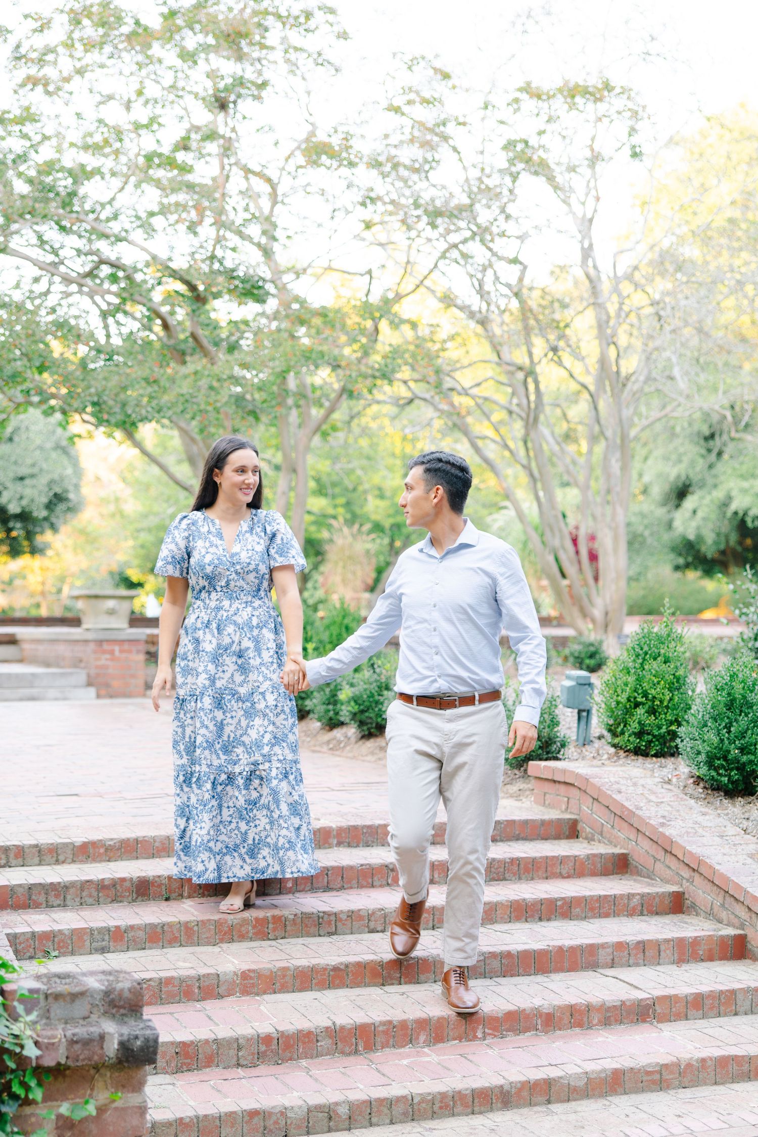 A couple holds hands while walking down brick steps in a lush garden, woman in blue floral dress.
