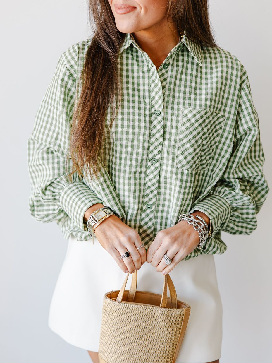 Woman wearing an oversized green gingham button-down shirt tucked into a white mini skirt, holding a straw tote.