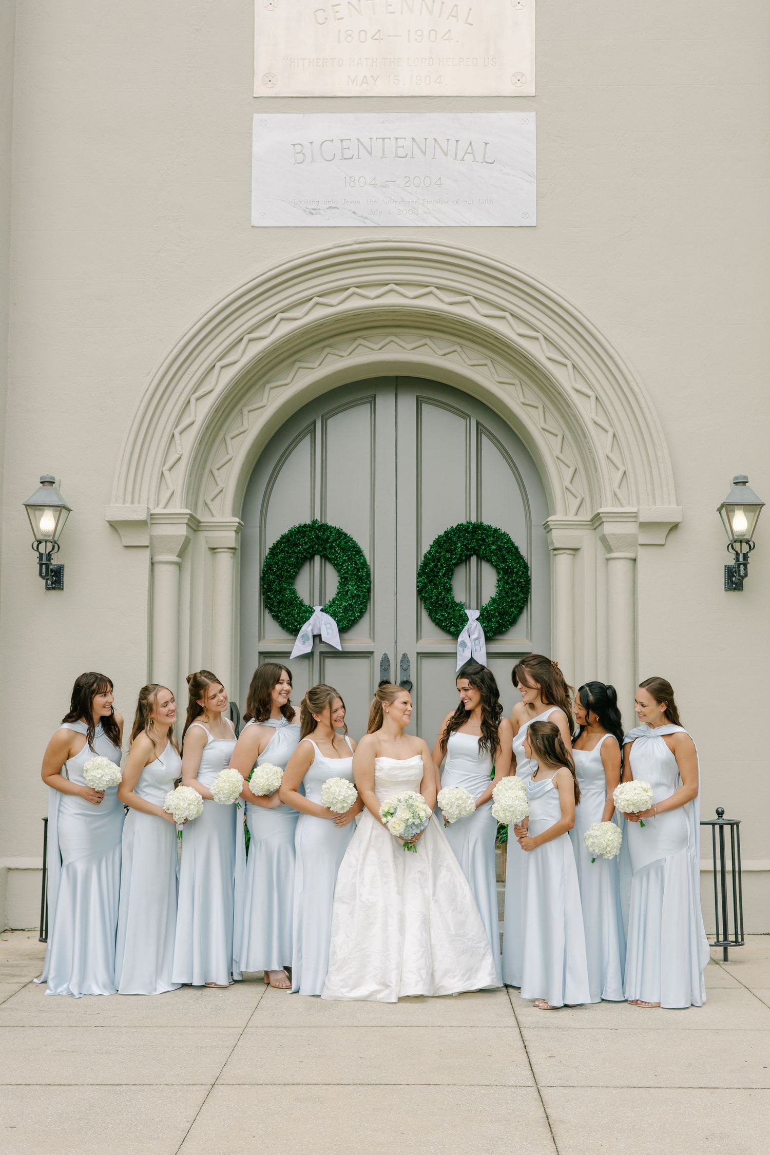 Bride and bridesmaids in light blue gowns pose with white bouquets before ornate church doors with green wreaths.