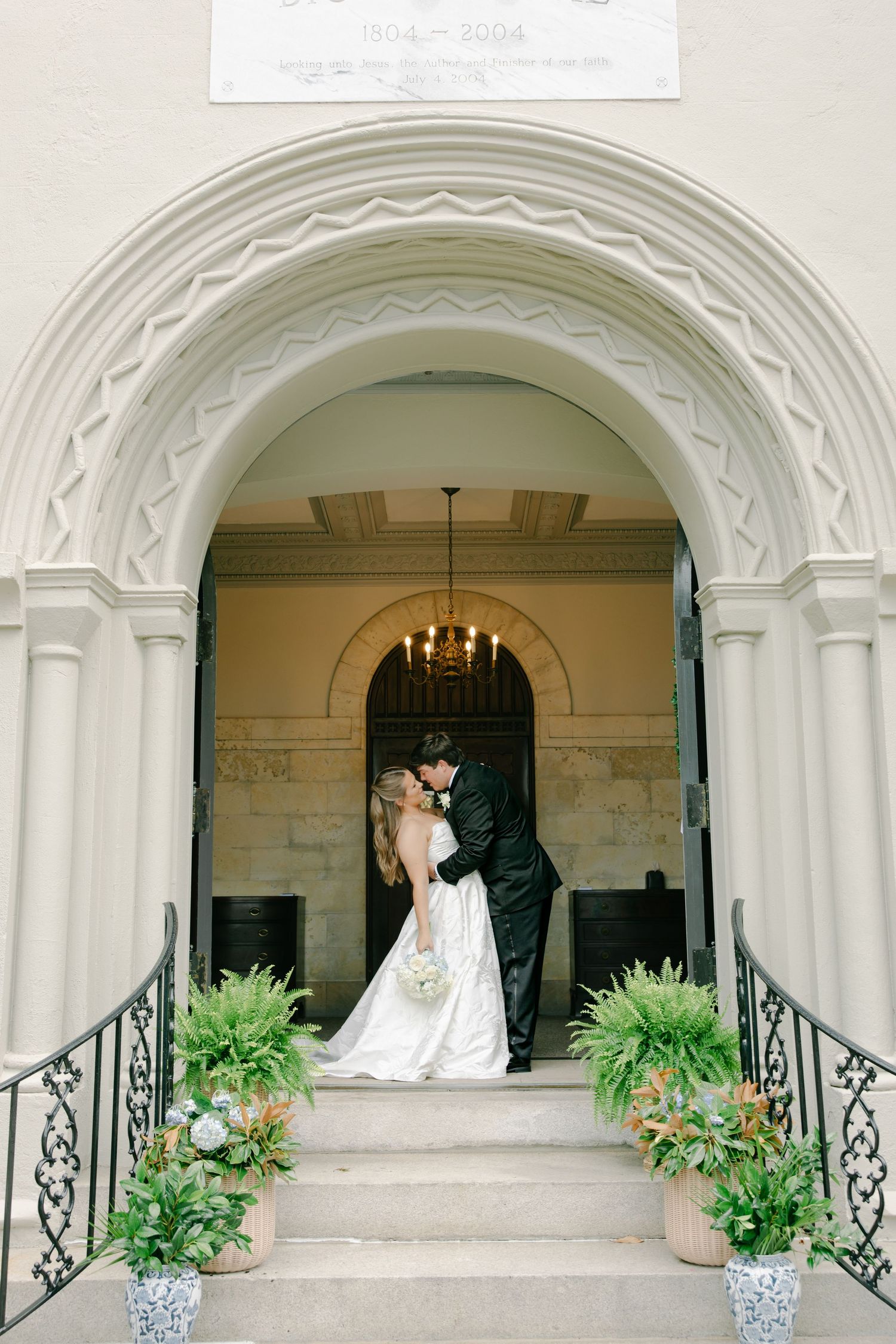 Bride and groom share a kiss under an ornate arched doorway adorned with ferns and floral arrangements on wedding day.