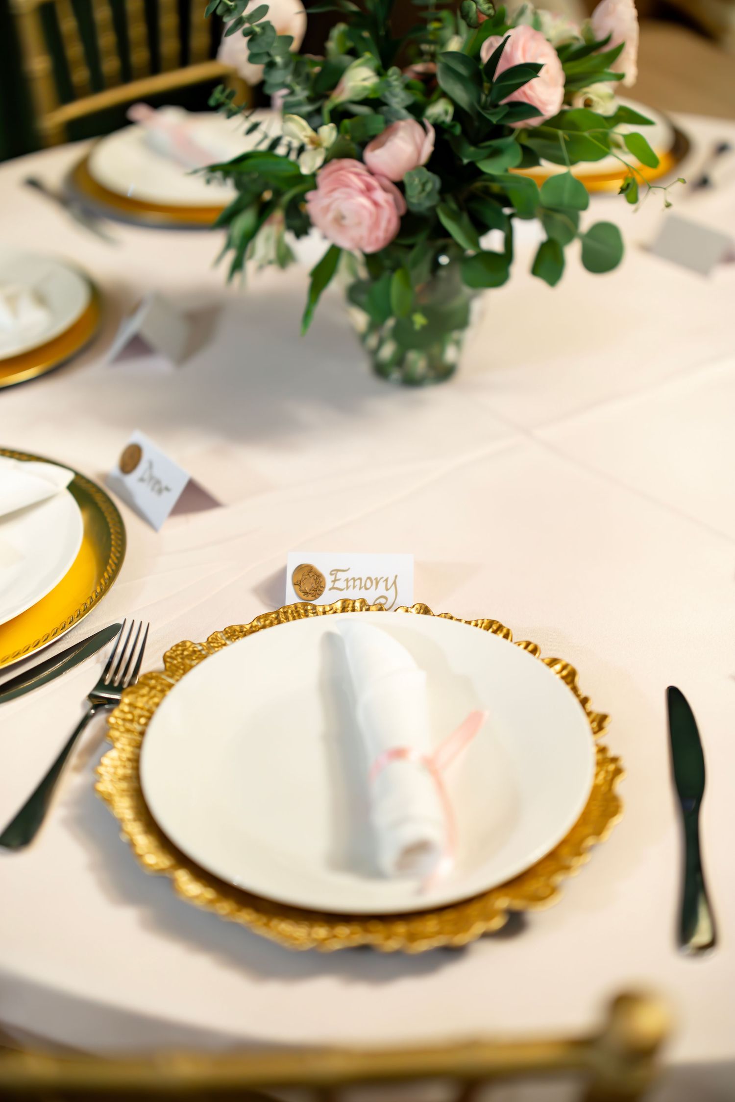 Gold charger plate with white napkin and place card at romantic wedding reception table with pink floral centerpiece.