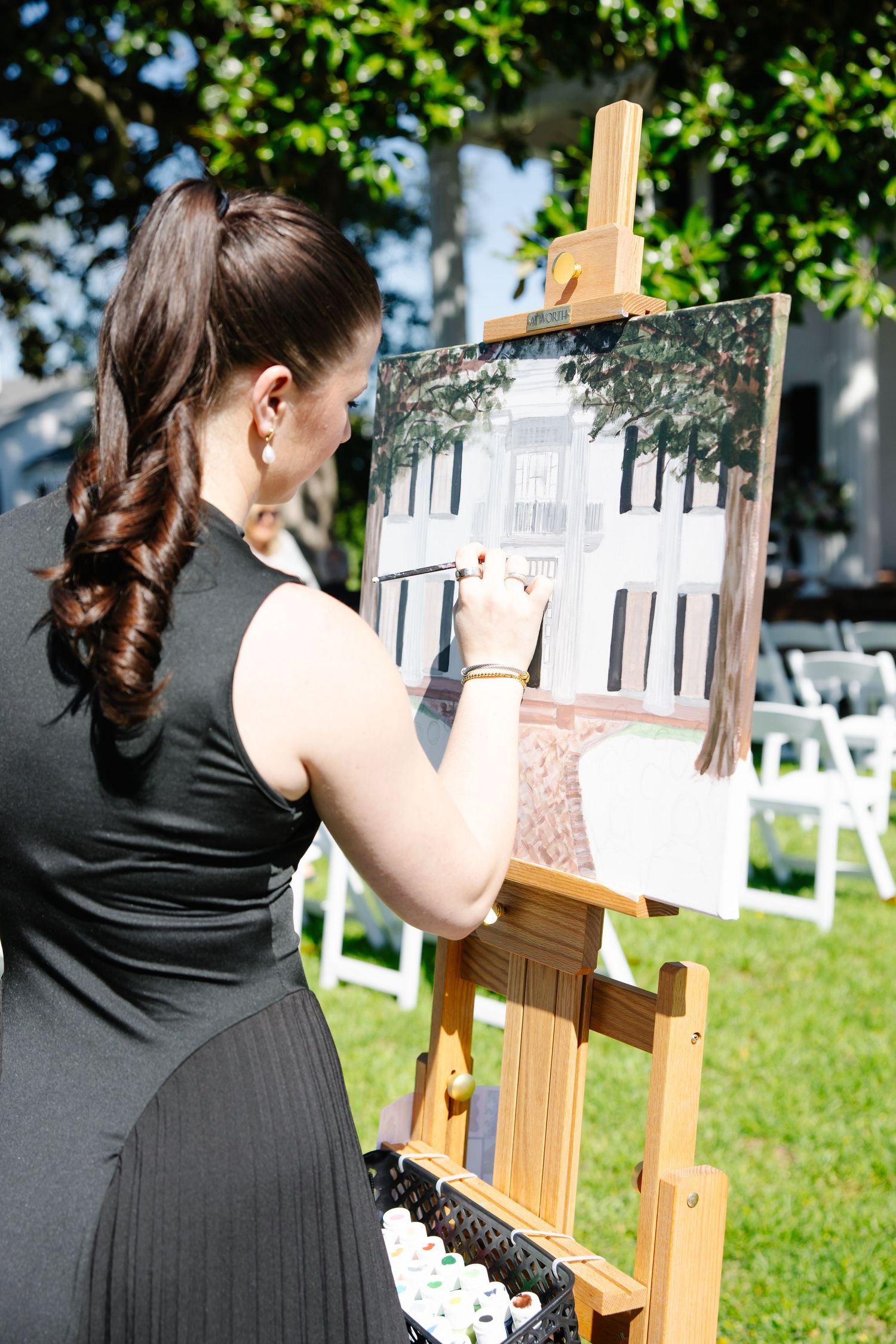 Artist in black dress painting wedding venue on easel during outdoor garden ceremony.