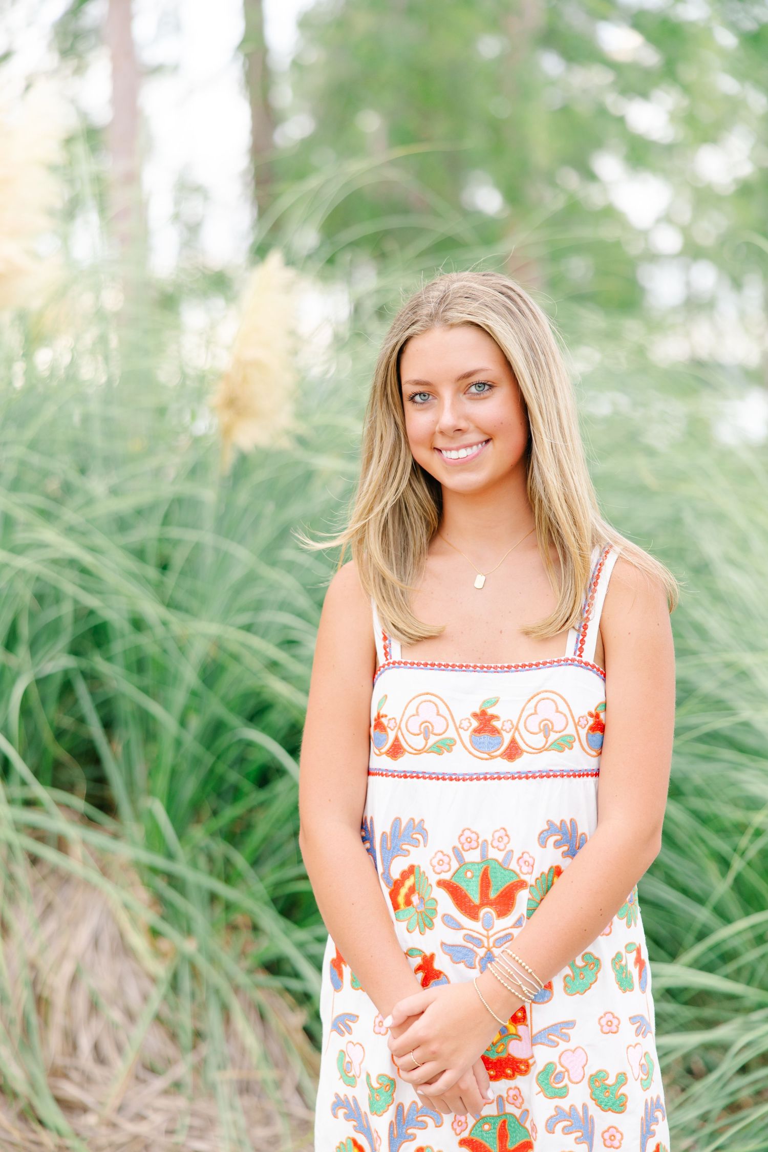 Smiling blonde teen in white floral embroidered sundress posing outdoors near tall ornamental grasses.