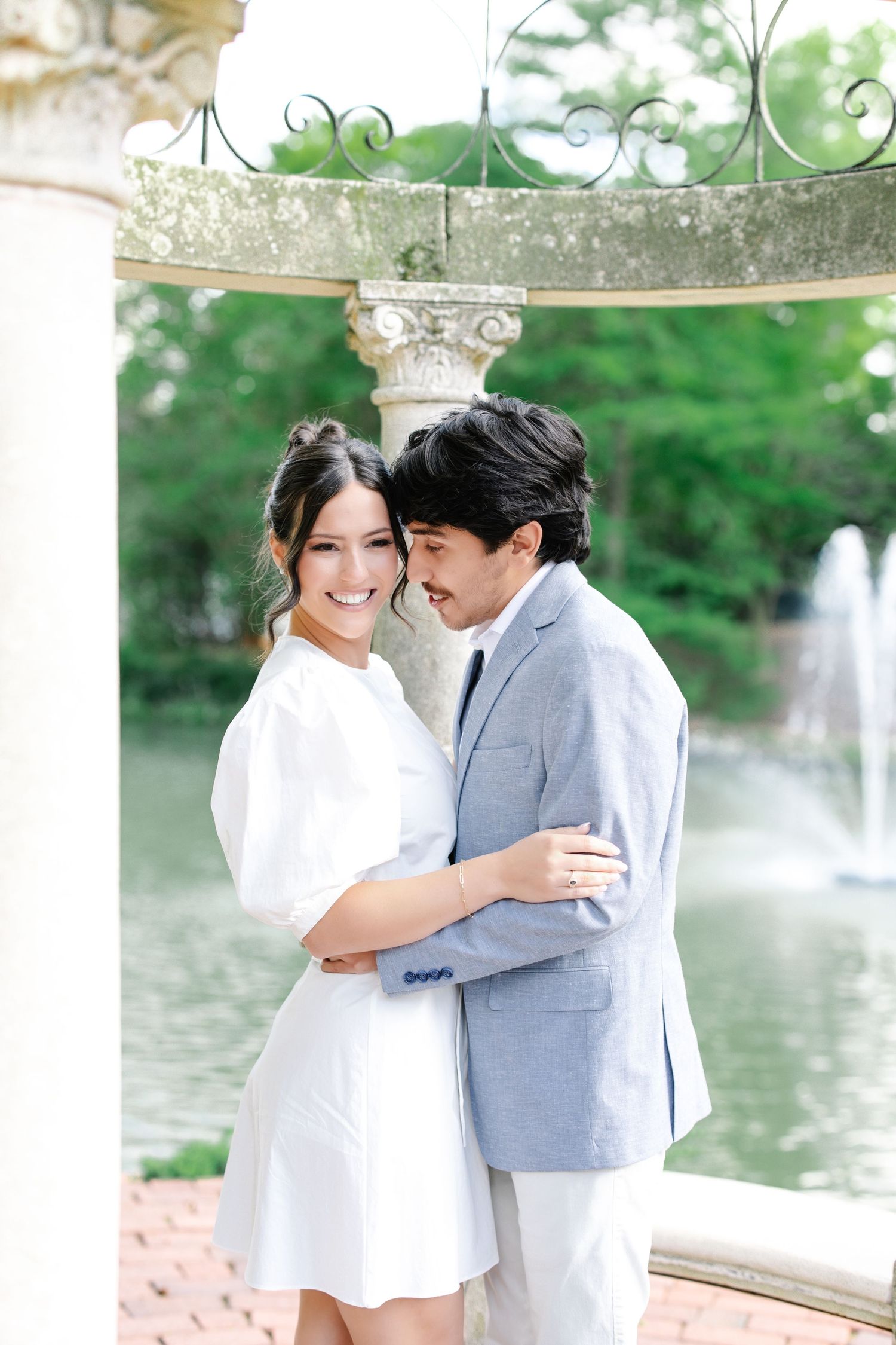 Smiling couple embracing near stone gazebo with fountain and green trees behind them.