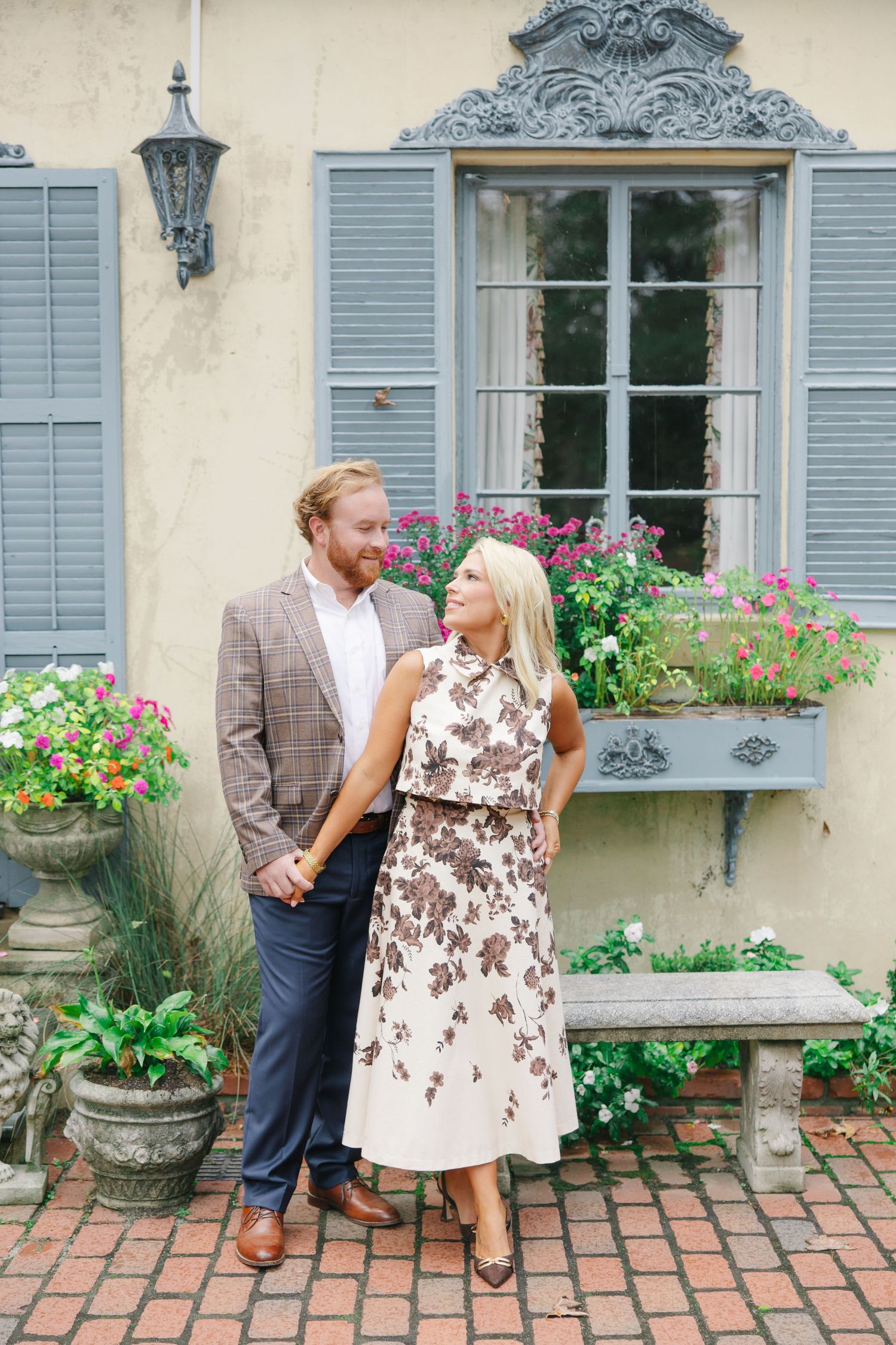 Couple in elegant attire standing before French-style yellow building with flower-filled window boxes.