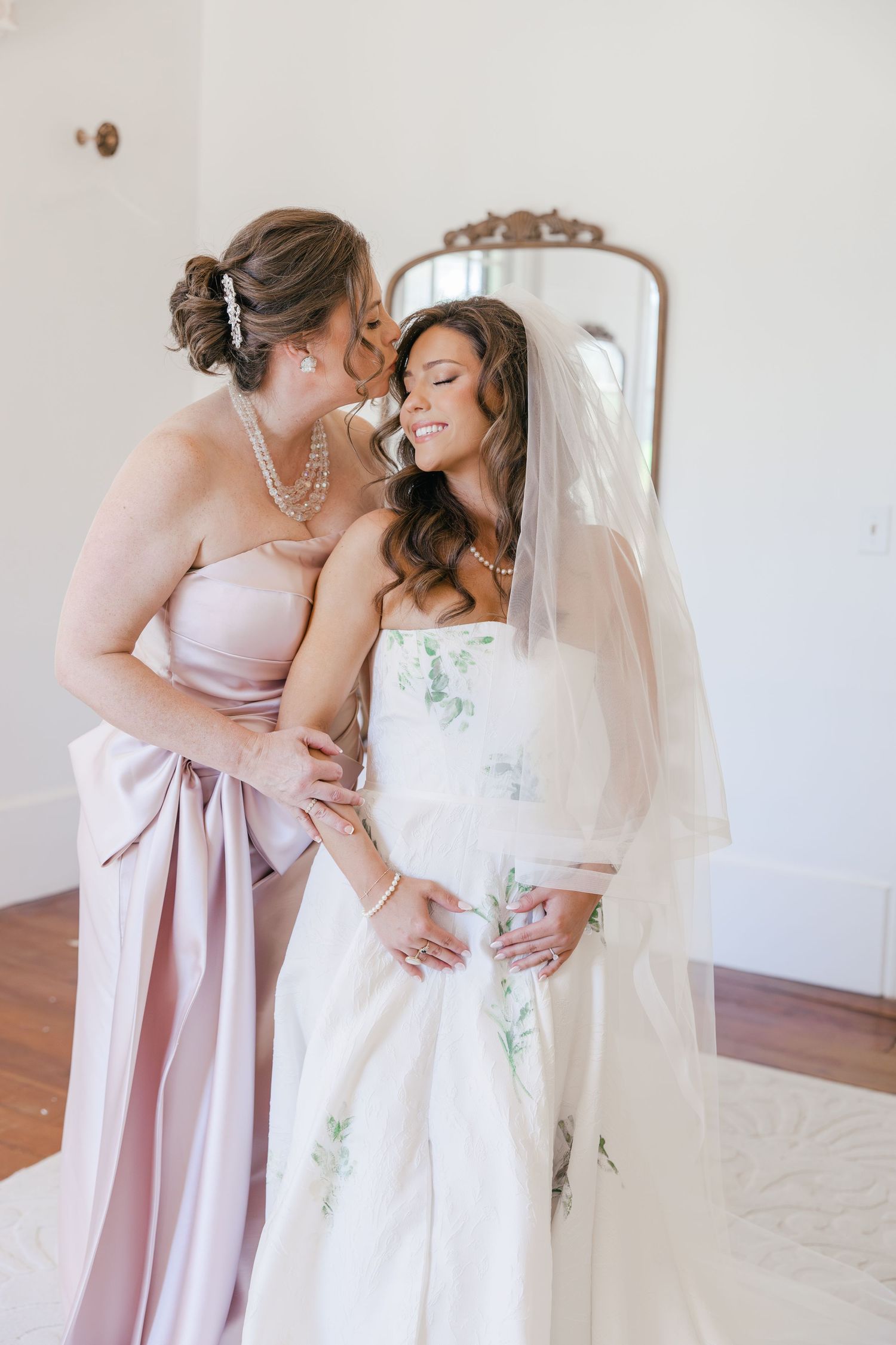 Mother of the bride whispering to smiling bride in floral strapless gown.