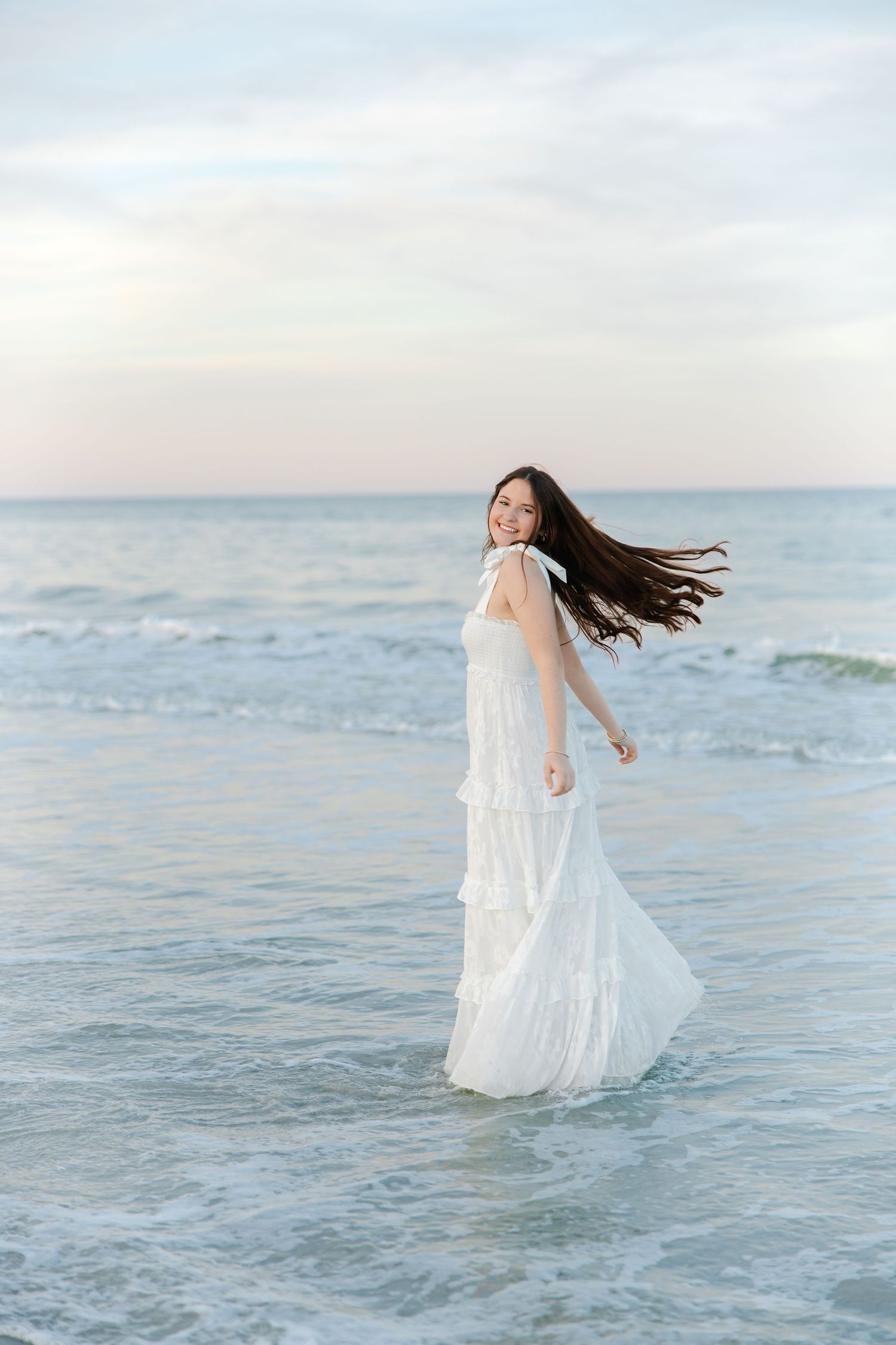 Young woman in white tiered maxi dress with hair flowing in wind, standing in ocean waves.