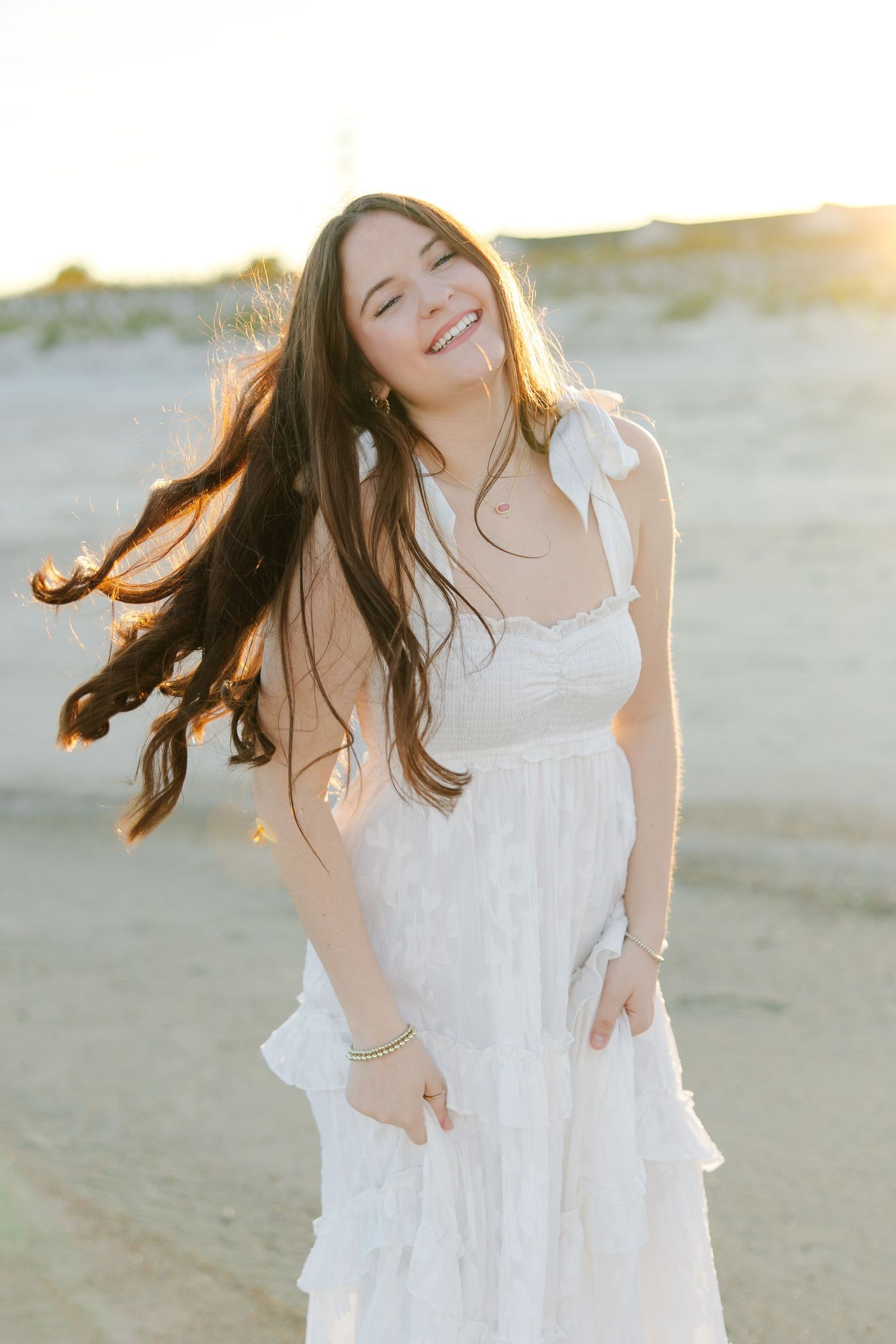 Young woman in white lace sundress laughs joyfully with hair blowing in golden sunset light.