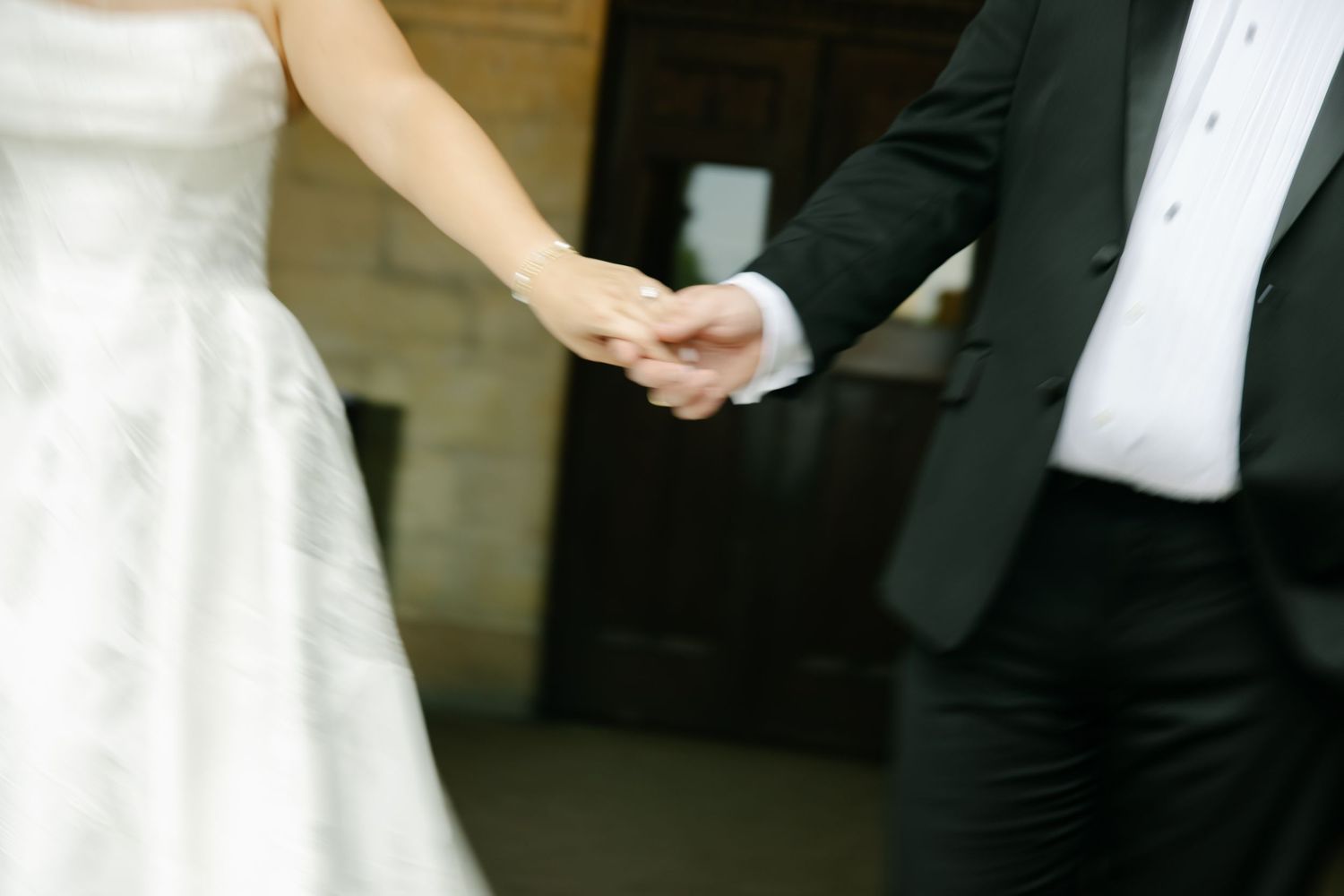 Bride and groom hold hands tenderly in close-up shot showing wedding attire details.